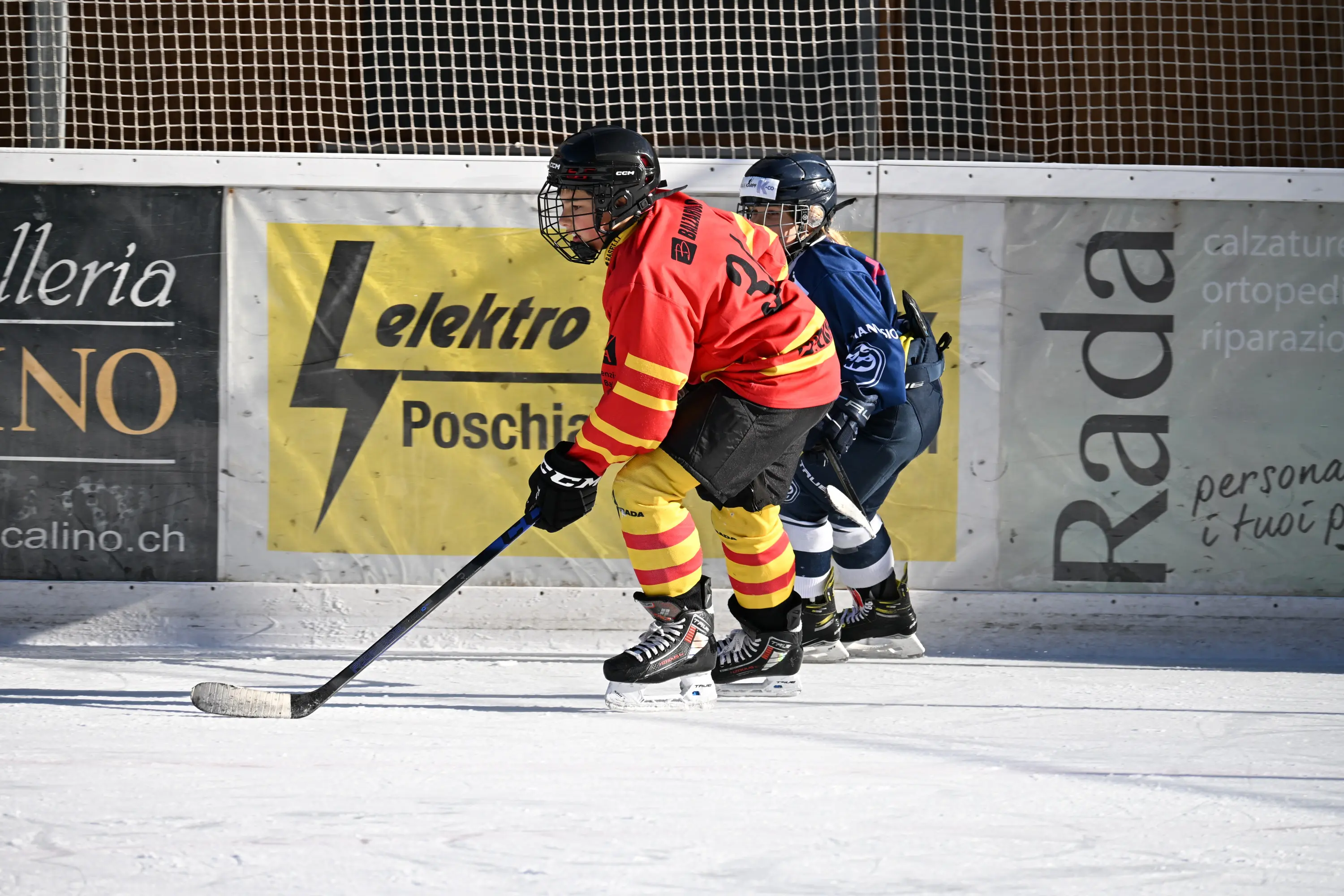 Giocatori di hockey su ghiaccio in azione. Un giocatore in maglia rossa e gialla è in movimento con un bastone da hockey, mentre un secondo giocatore in mag
