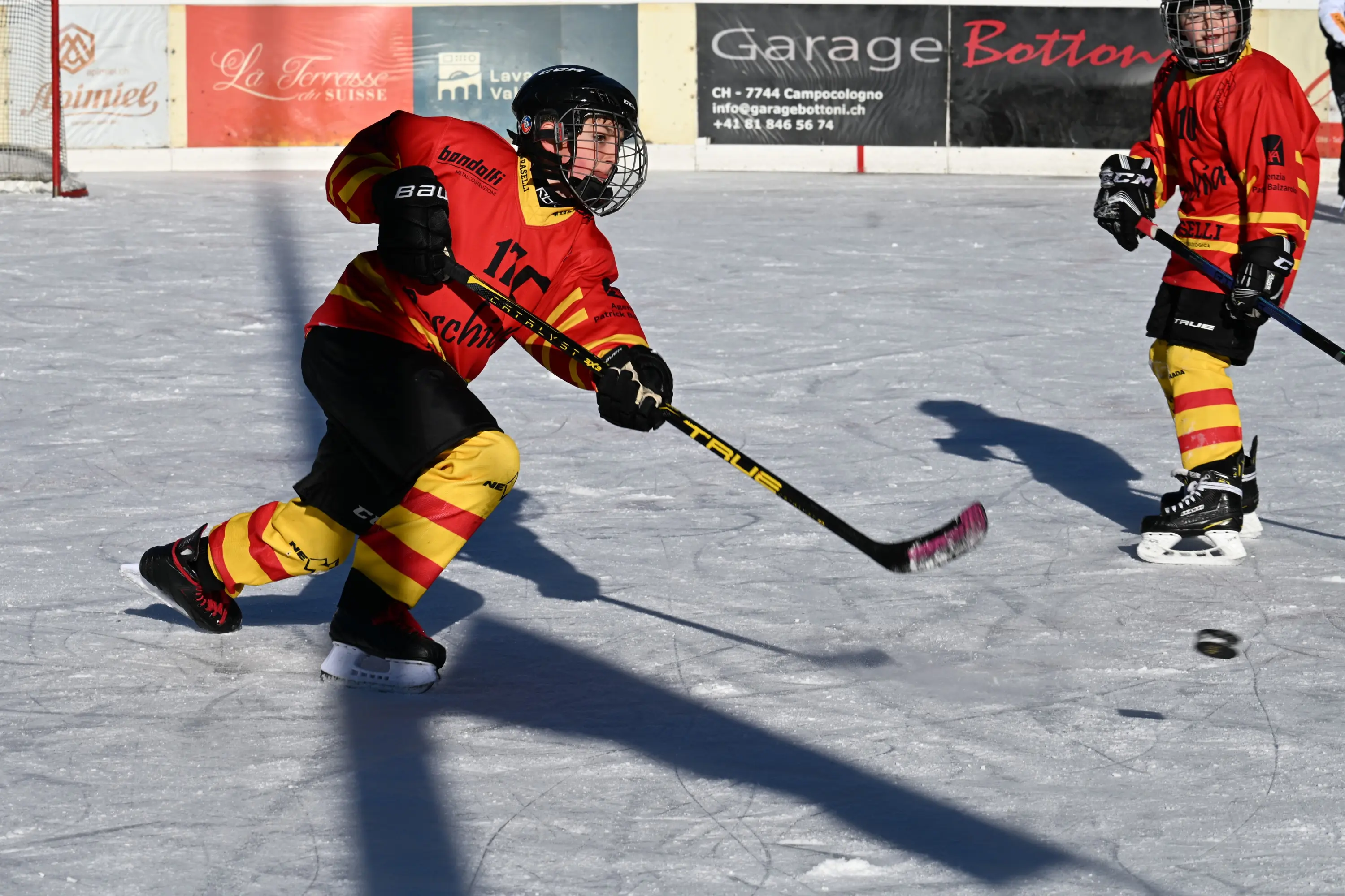 Due giovani giocatori di hockey su ghiaccio indossano divise rosse e gialle mentre si allenano su un campo ghiacciato. Uno dei ragazzi sta colpendo il disco