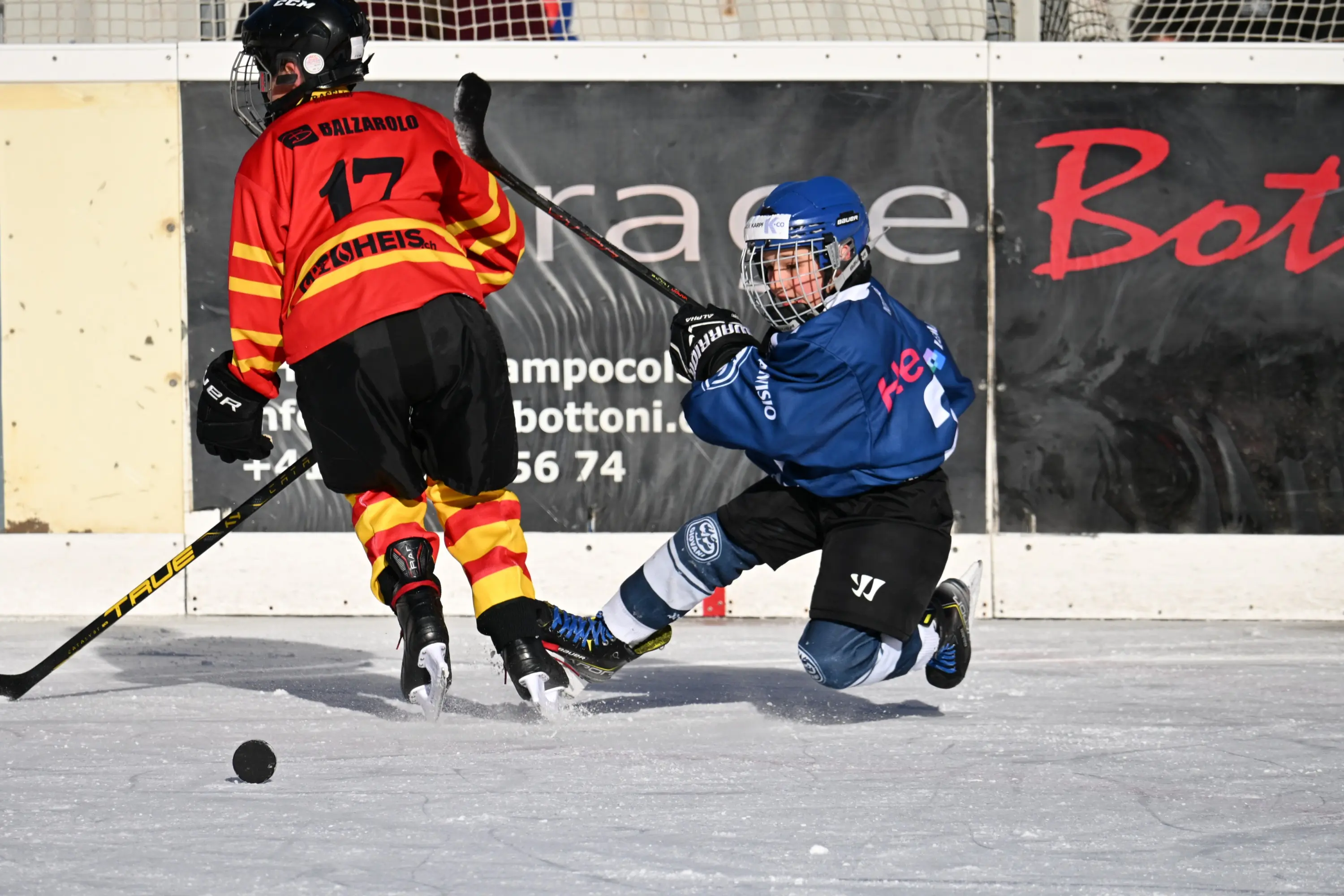 Giocatori di hockey su ghiaccio in azione, uno in maglia blu cade mentre cerca di colpire il disco, l'altro indossa una maglia rossa e gialla