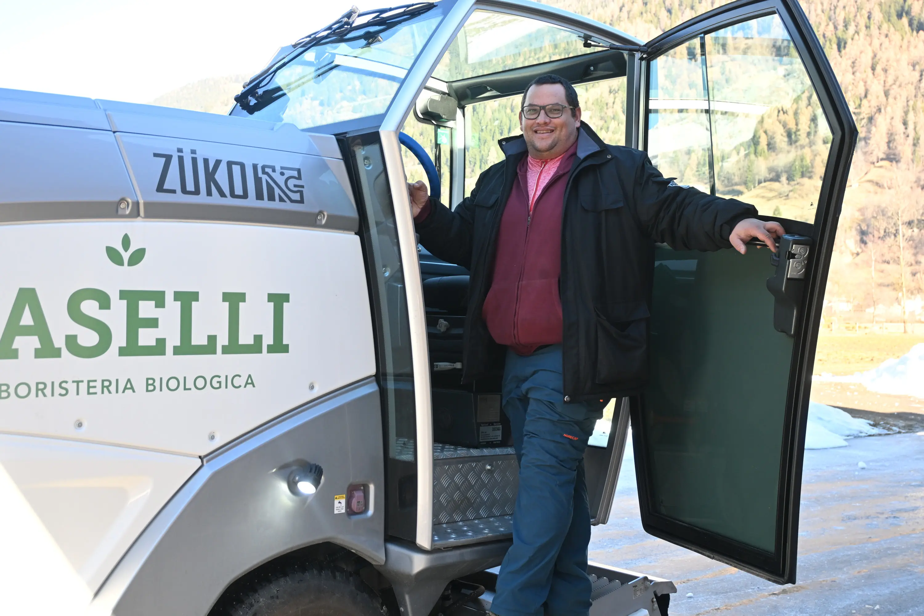 Uomo sorridente in piedi accanto a un veicolo all'aperto, con porte aperte. Indossa un giubbotto scuro e una maglietta rossa, mentre si