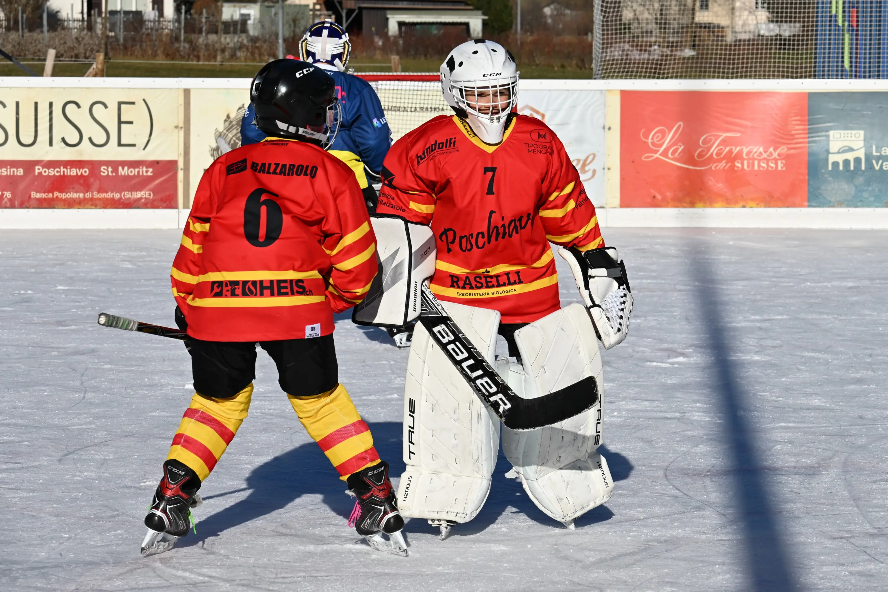 Due giocatori di hockey su ghiaccio con maglie rosse e gialle. Uno indossa un casco e guanti da portiere, mentre l'altro, senza casco, si prepara