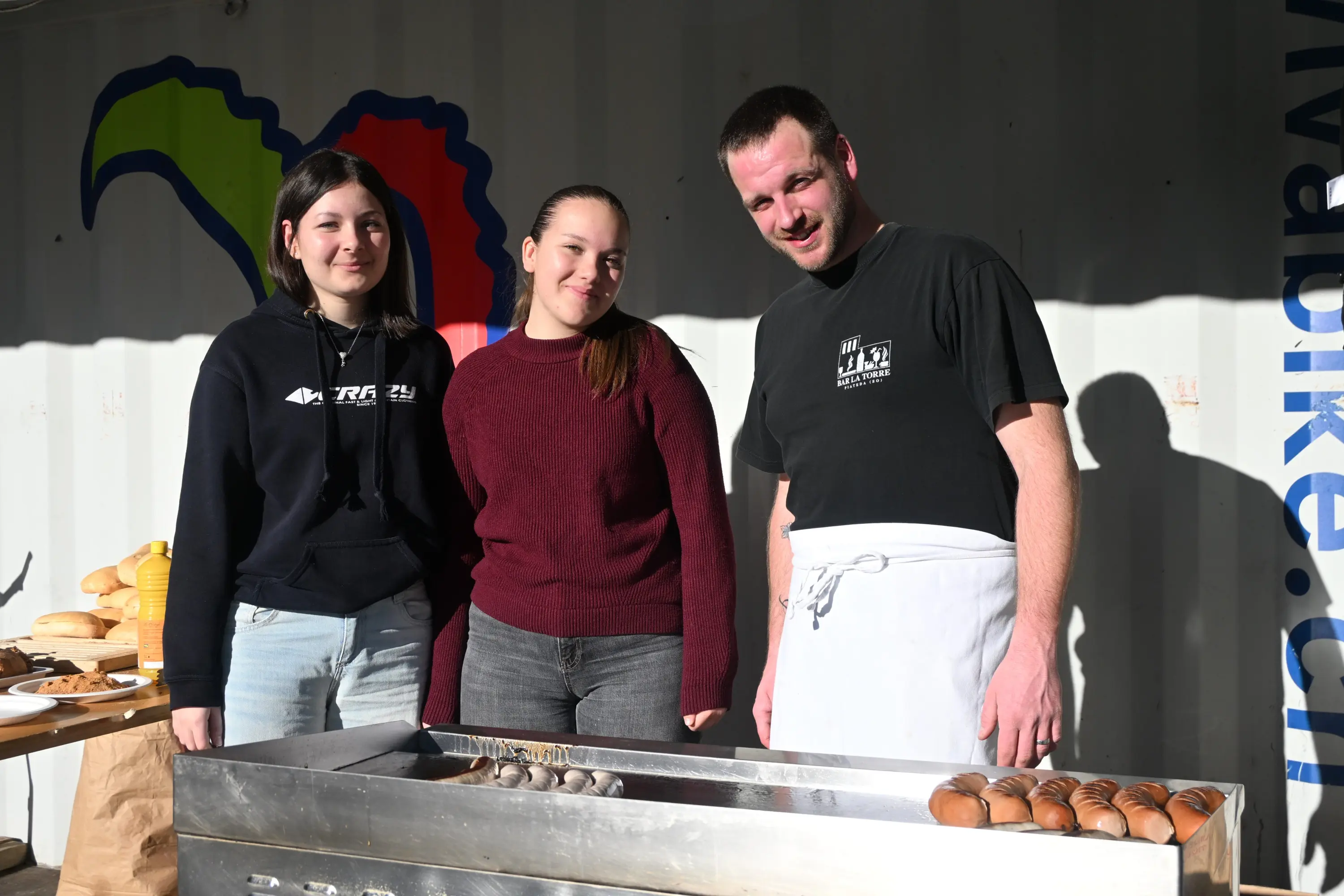 Tre persone sorridono davanti a un forno. Due ragazze indossano maglioni, mentre un uomo in camicia da cuoco è dietro il banco di lavoro. Sullo sf