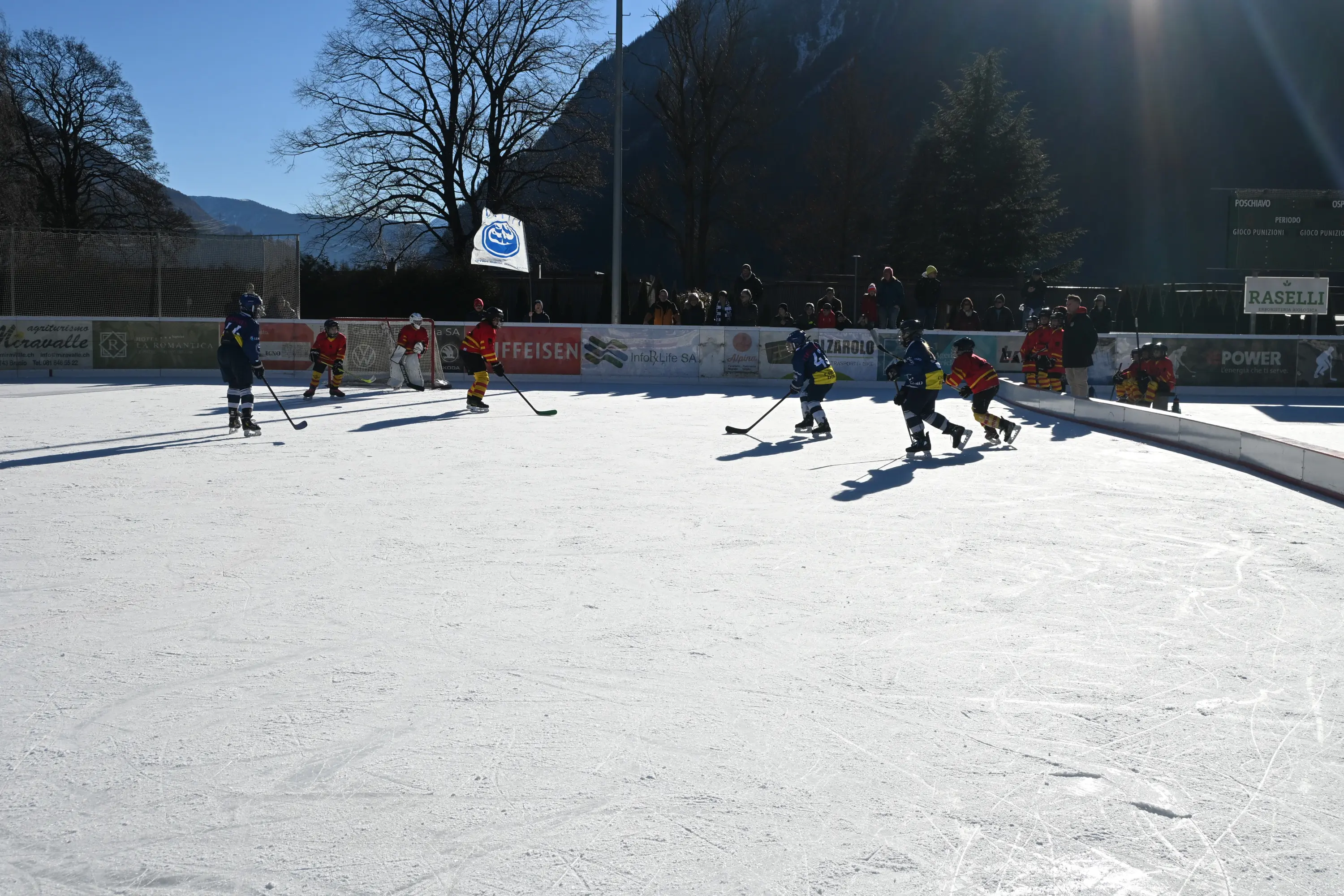 Giocatori di hockey su ghiaccio si sfidano su un campo all'aperto, circondati da alberi e spettatori. È una giornata di sole con un cielo chiaro.