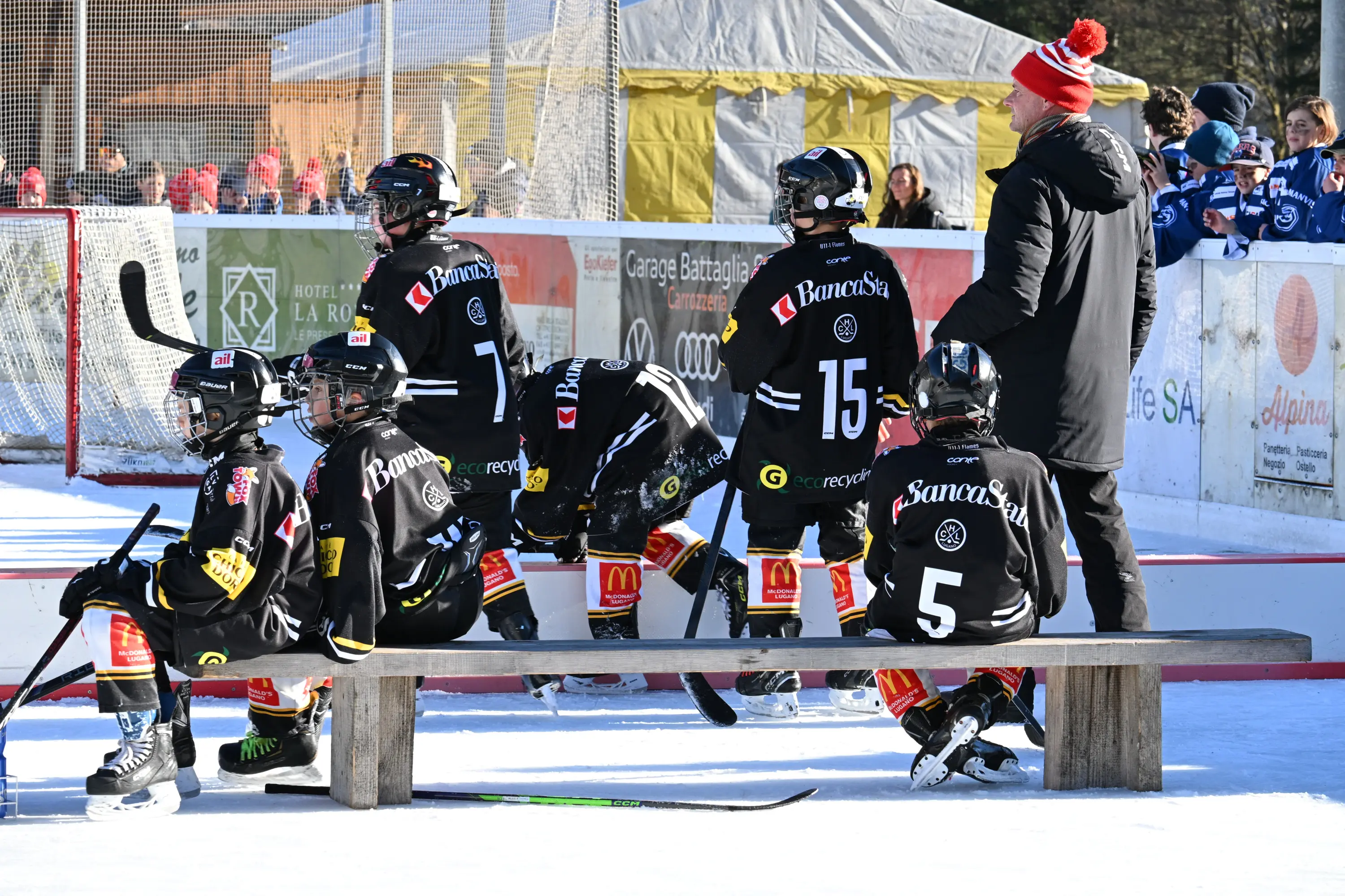 Un gruppo di giovani giocatori di hockey su ghiaccio indossa maglie nere e si concentra durante una pausa. Sono seduti su una panchina all'aperto, con un allenatore che