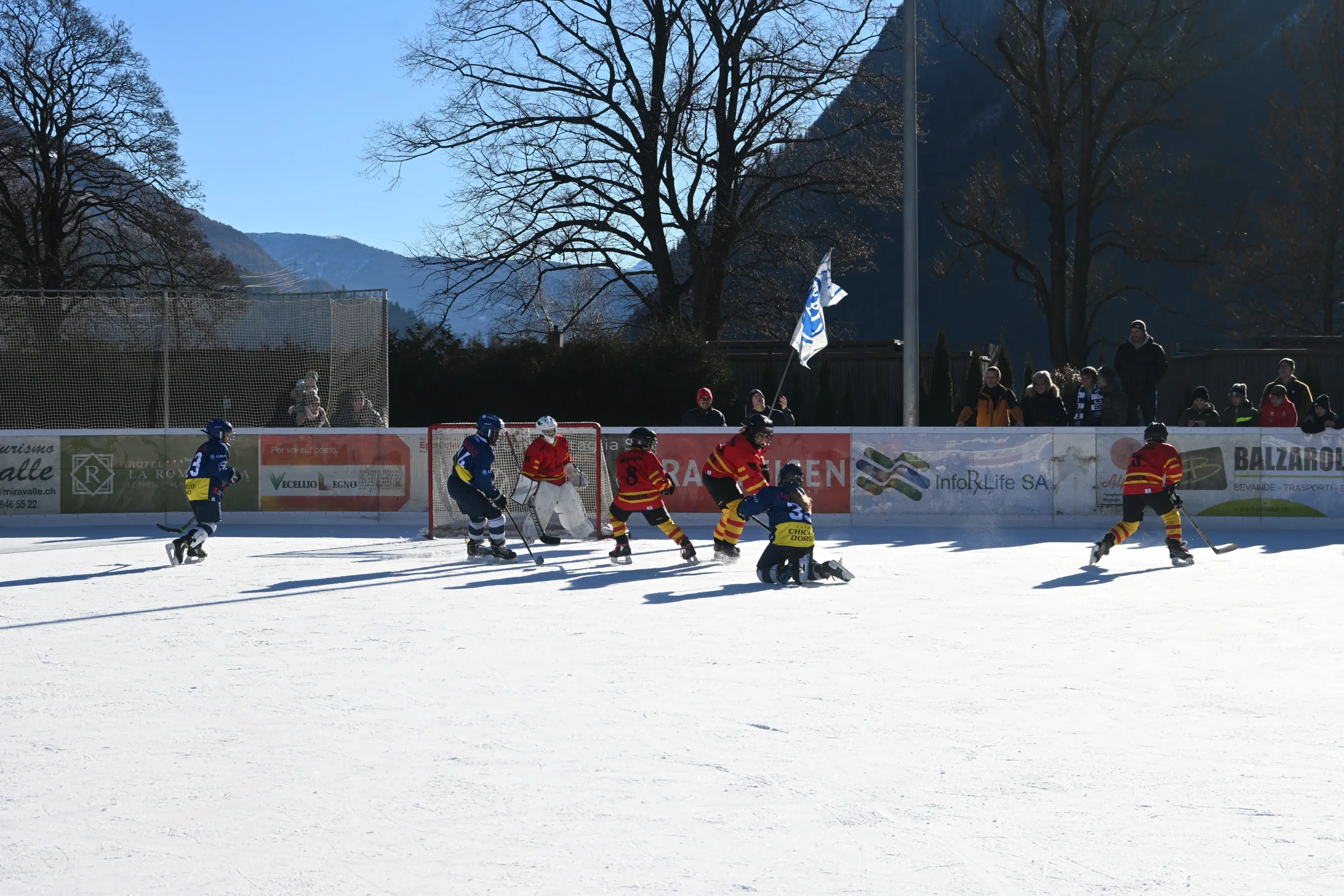 Giocatori di hockey su ghiaccio in azione su un campo ghiacciato all'aperto. Alcuni bambini indossano maglie colorate mentre un portiere è in posizione. Sul