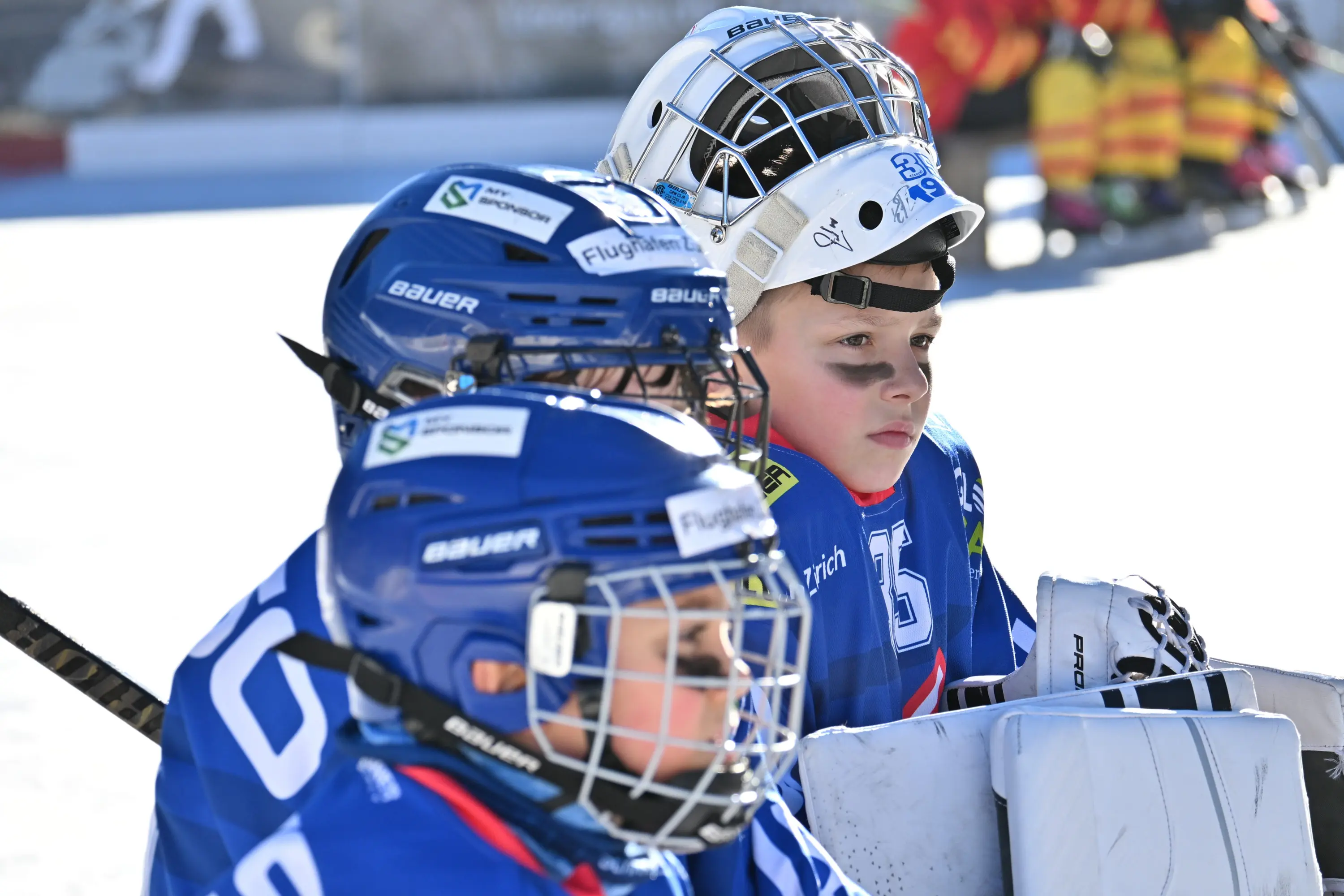 Giovani giocatori di hockey su ghiaccio in uniforme blu, concentrati e in attesa. Hanno caschi bianchi e protezioni, con uno che guarda lontano. Sullo sf