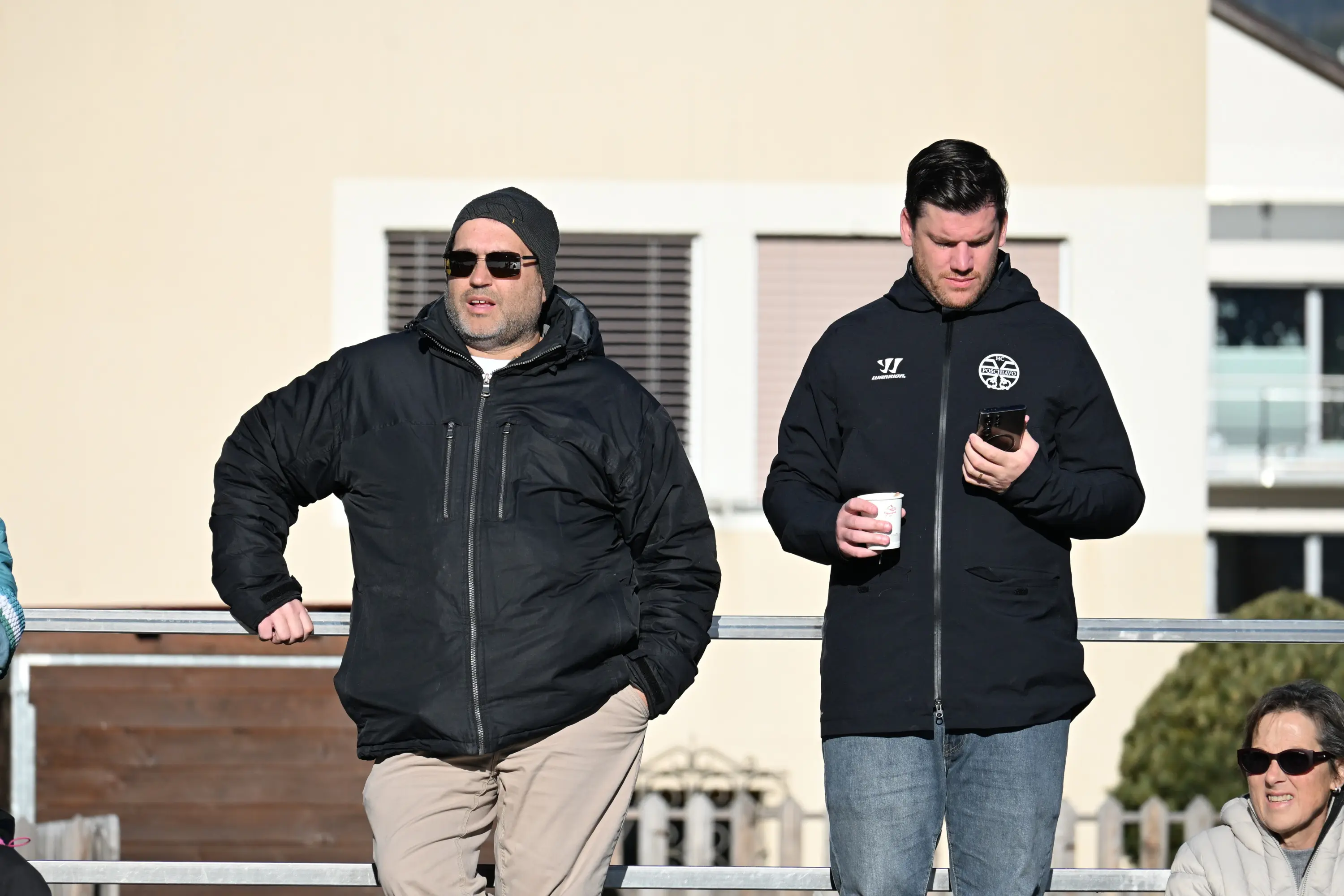 Due uomini in abbigliamento invernale stanno in piedi su una balconata. Uno indossa un berretto e occhiali da sole, mentre l'altro ha in mano un cellul