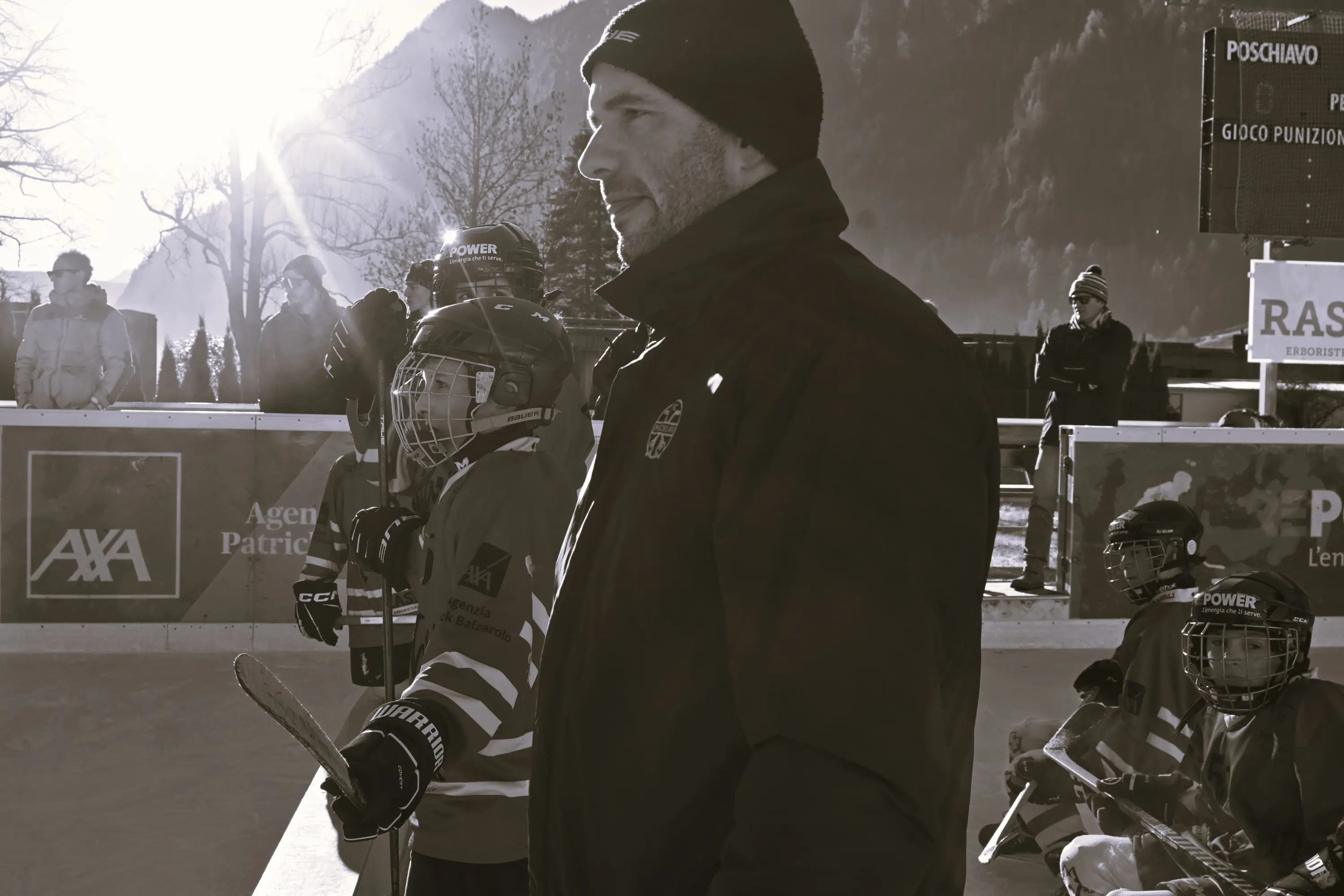 Un allenatore di hockey su ghiaccio osserva i giovani giocatori mentre si preparano per una partita all'aperto, con montagne sullo sfondo e il sole che illumina la scena.