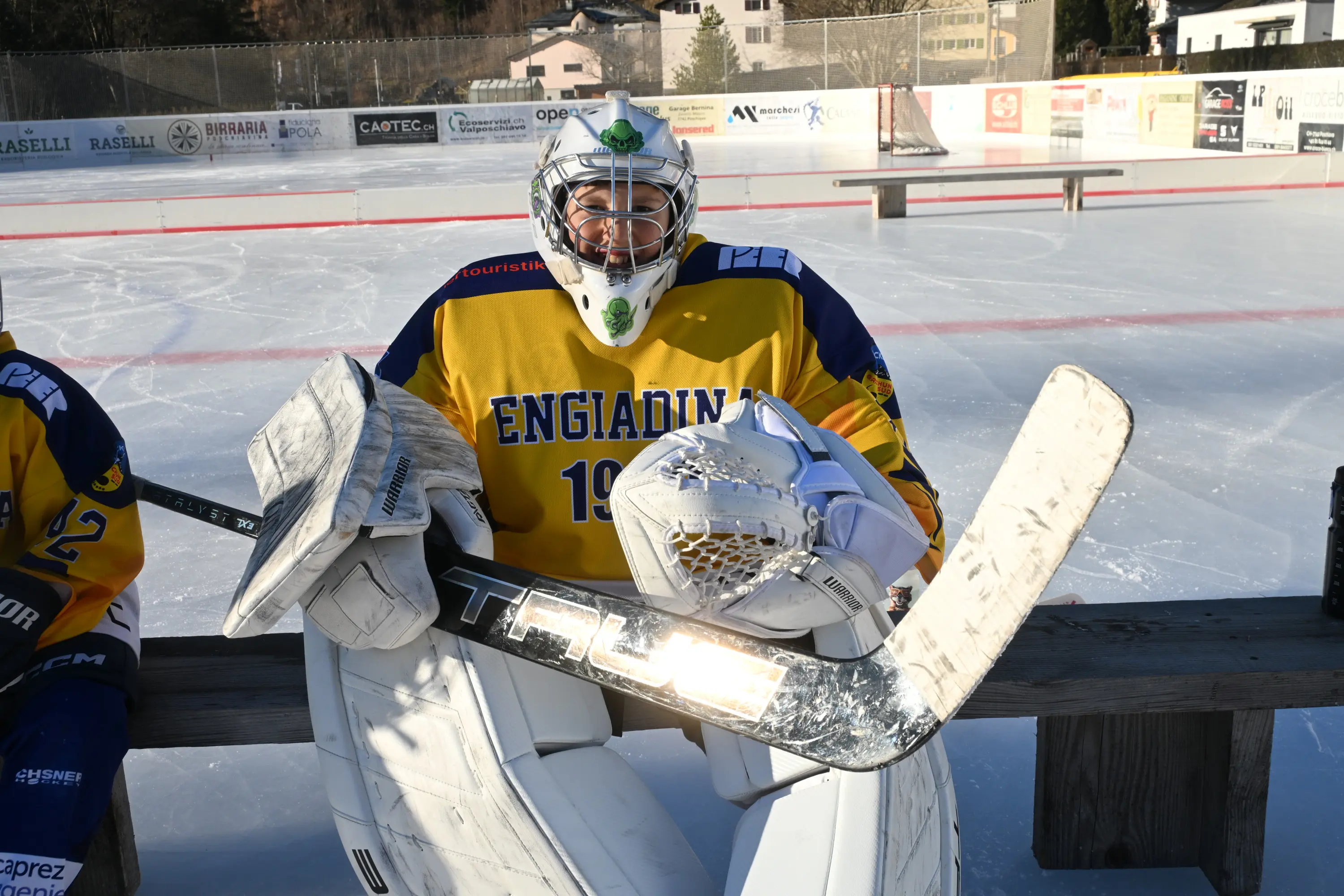 Un portiere di hockey su ghiaccio indossa un'uniforme gialla con la scritta "ENGADIN" e il numero 19. Si trova su una panchina vicino a