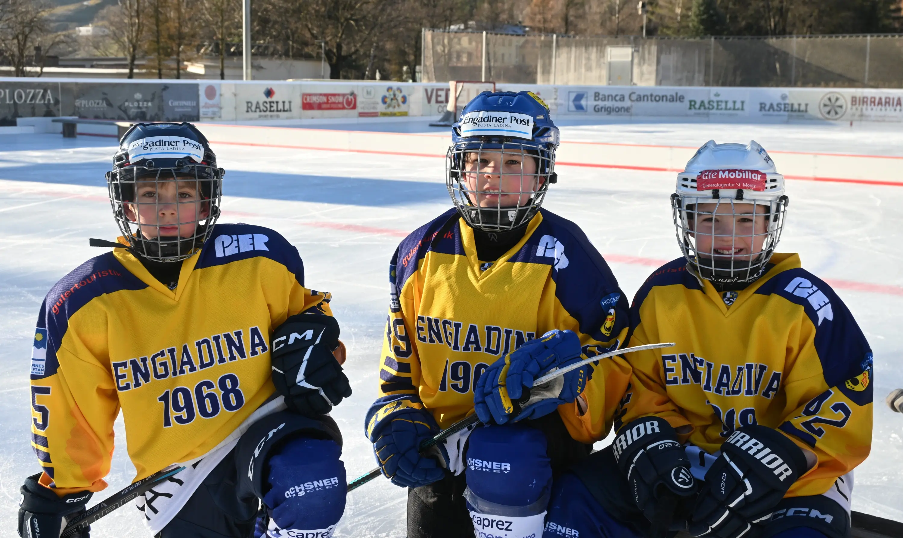 Tre giovani giocatori di hockey su ghiaccio indossano divise gialle con scritte blu. Sono in una pista di ghiaccio all'aperto, sorridendo e posando per