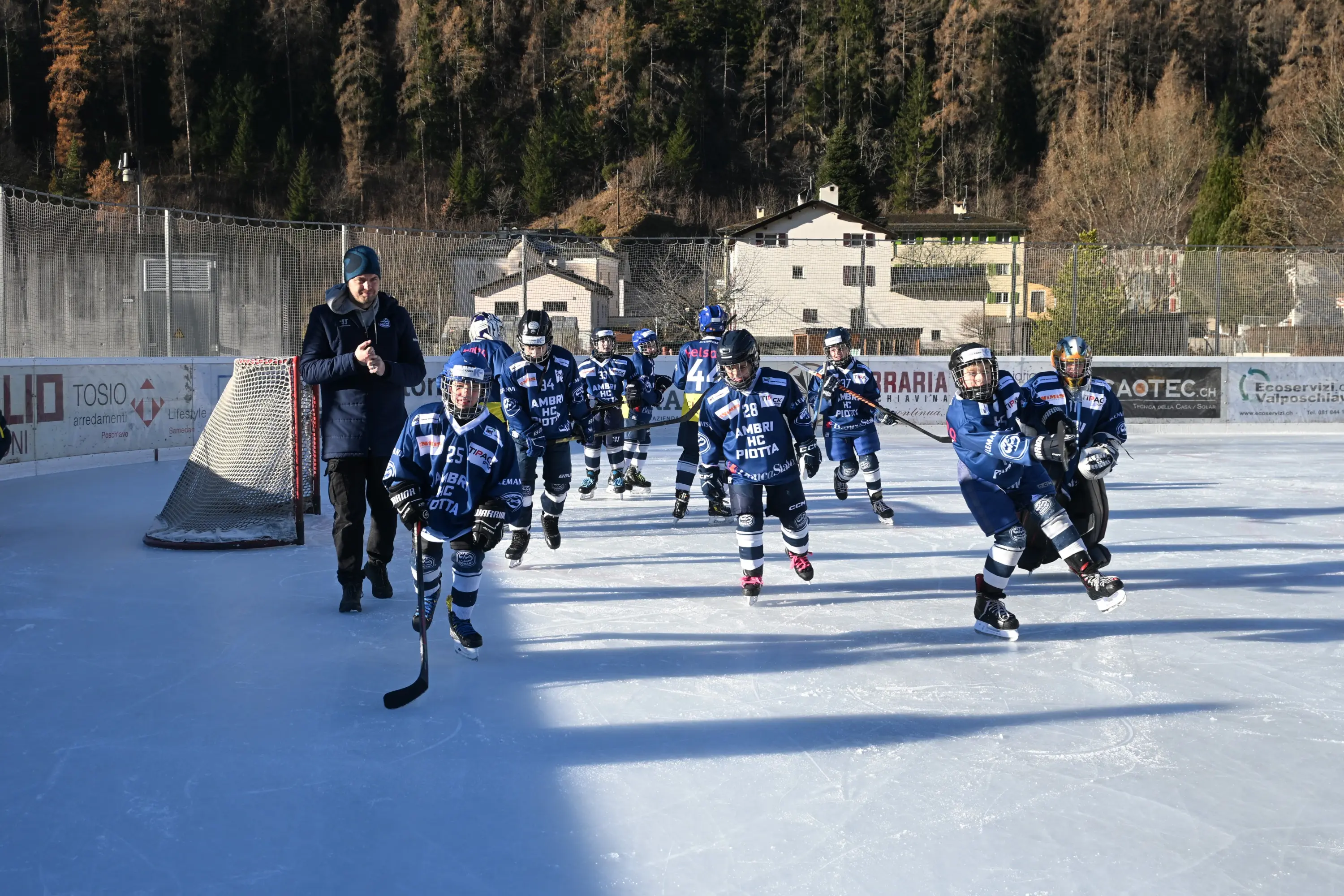 Una squadra di giovani hockeisti in divise blu si allena sul ghiaccio. Alcuni bambini pattinano mentre un allenatore osserva e guida. Sullo sfondo si ved