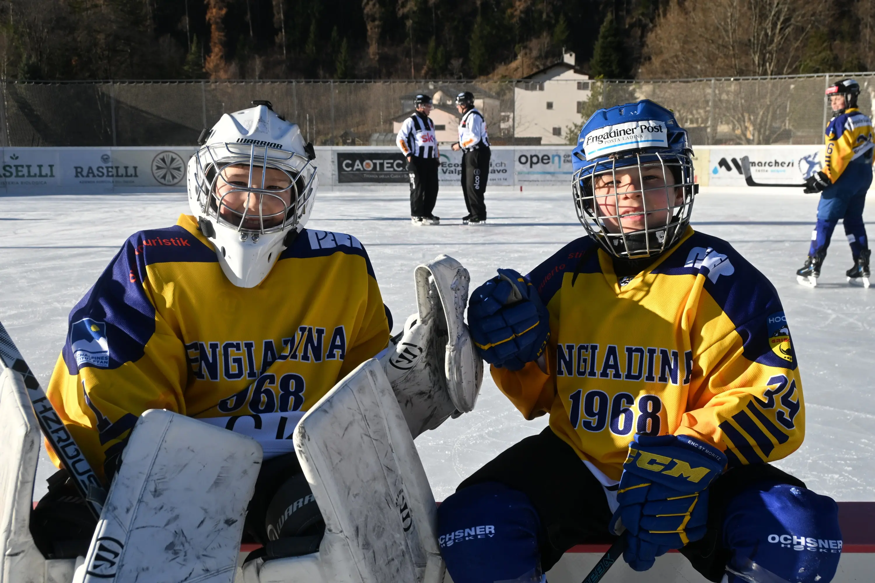 Due giovani hockeisti sorridenti indossano maglie gialle e blu con il logo "Engiadina 1968". Sono in una pista di ghiaccio, circondati