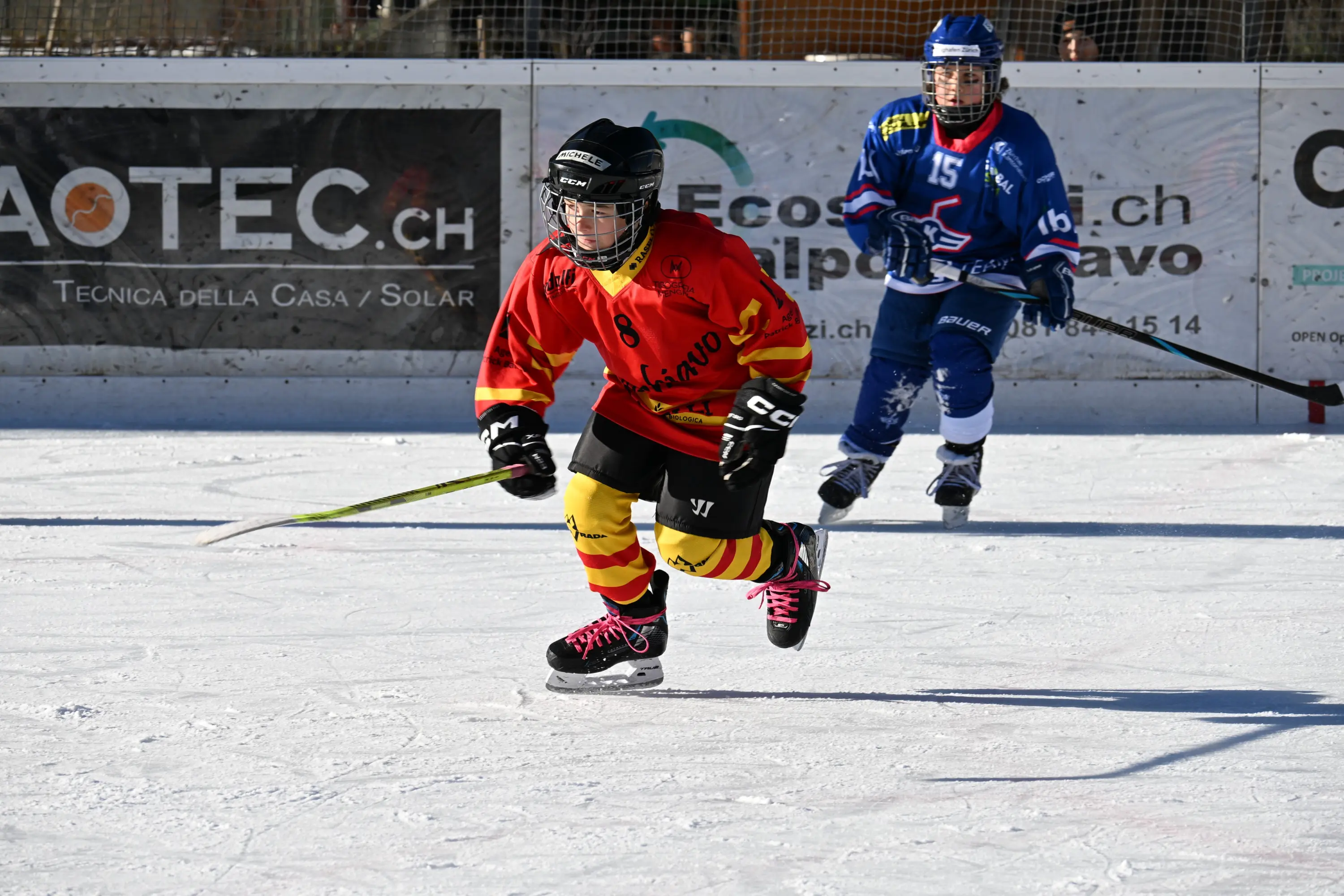 Giocatore di hockey su ghiaccio in uniforme rosso e giallo, in movimento sul ghiaccio. Sullo sfondo, un altro giocatore in uniforme blu.