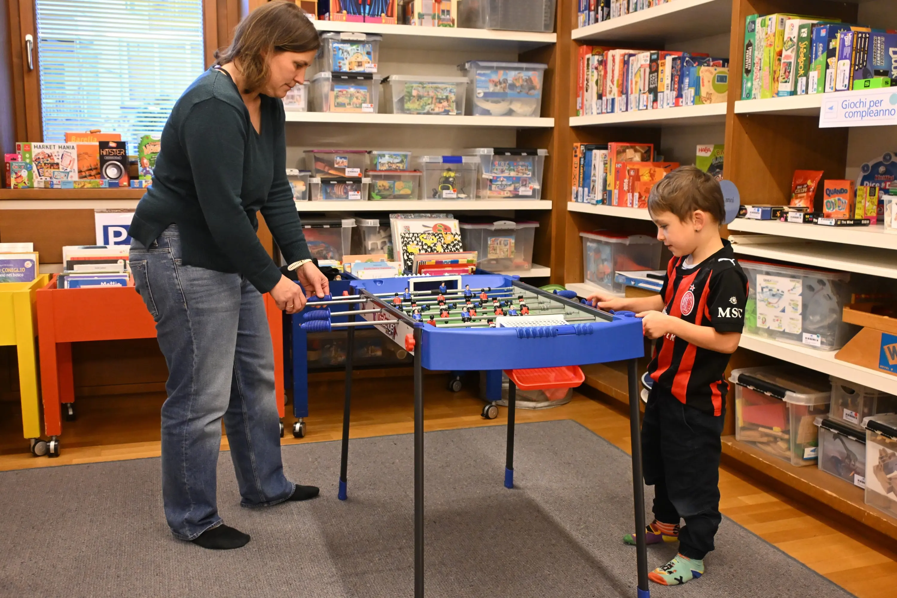 Una donna gioca a biliardino con un bambino in una biblioteca. Sullo sfondo si vedono scaffali pieni di libri e giochi.