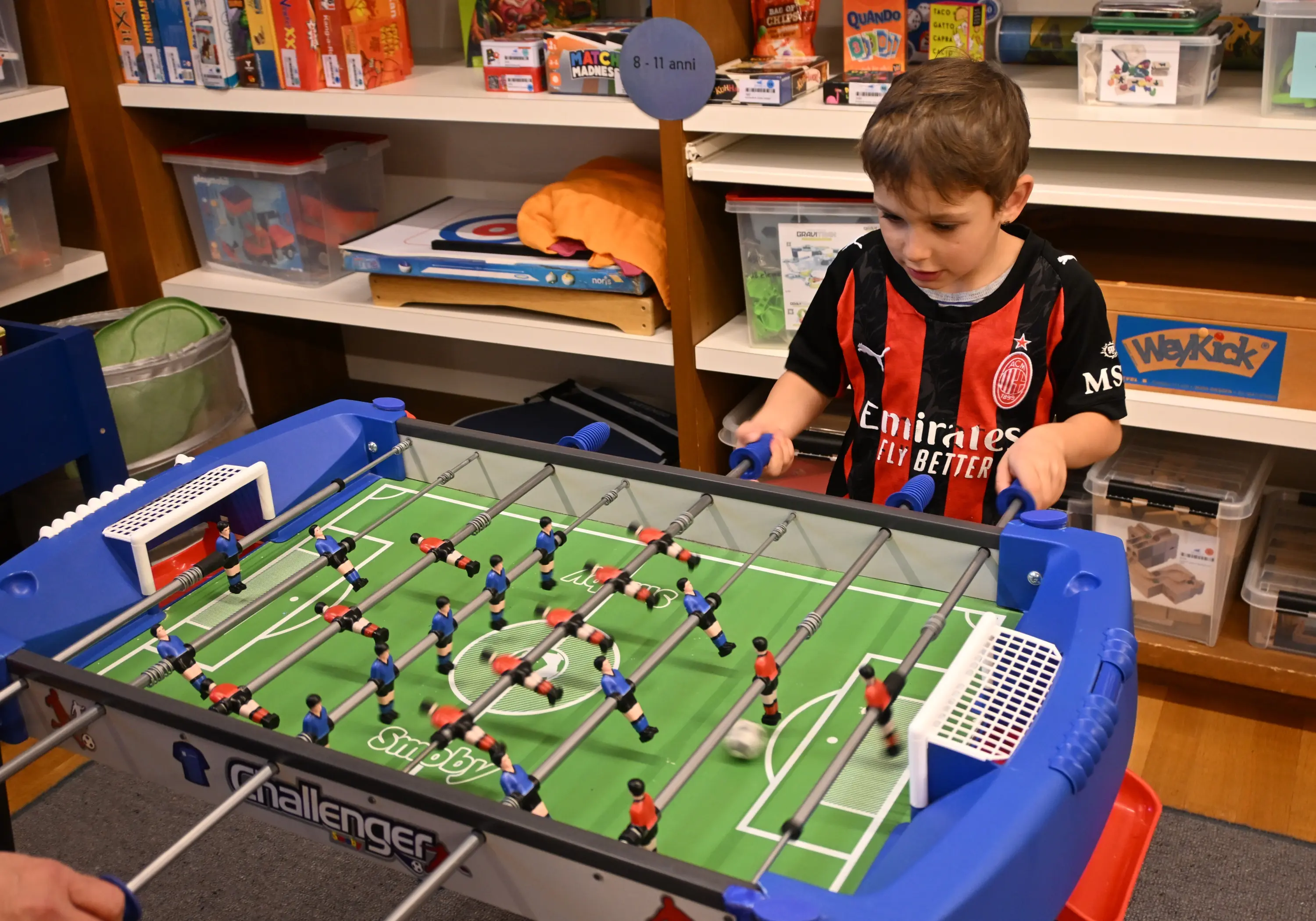 Un bambino gioca a biliardino, concentrato mentre muove le stecche. Indossa una maglietta rossa e nera. Sullo sfondo si vedono scaffali pieni