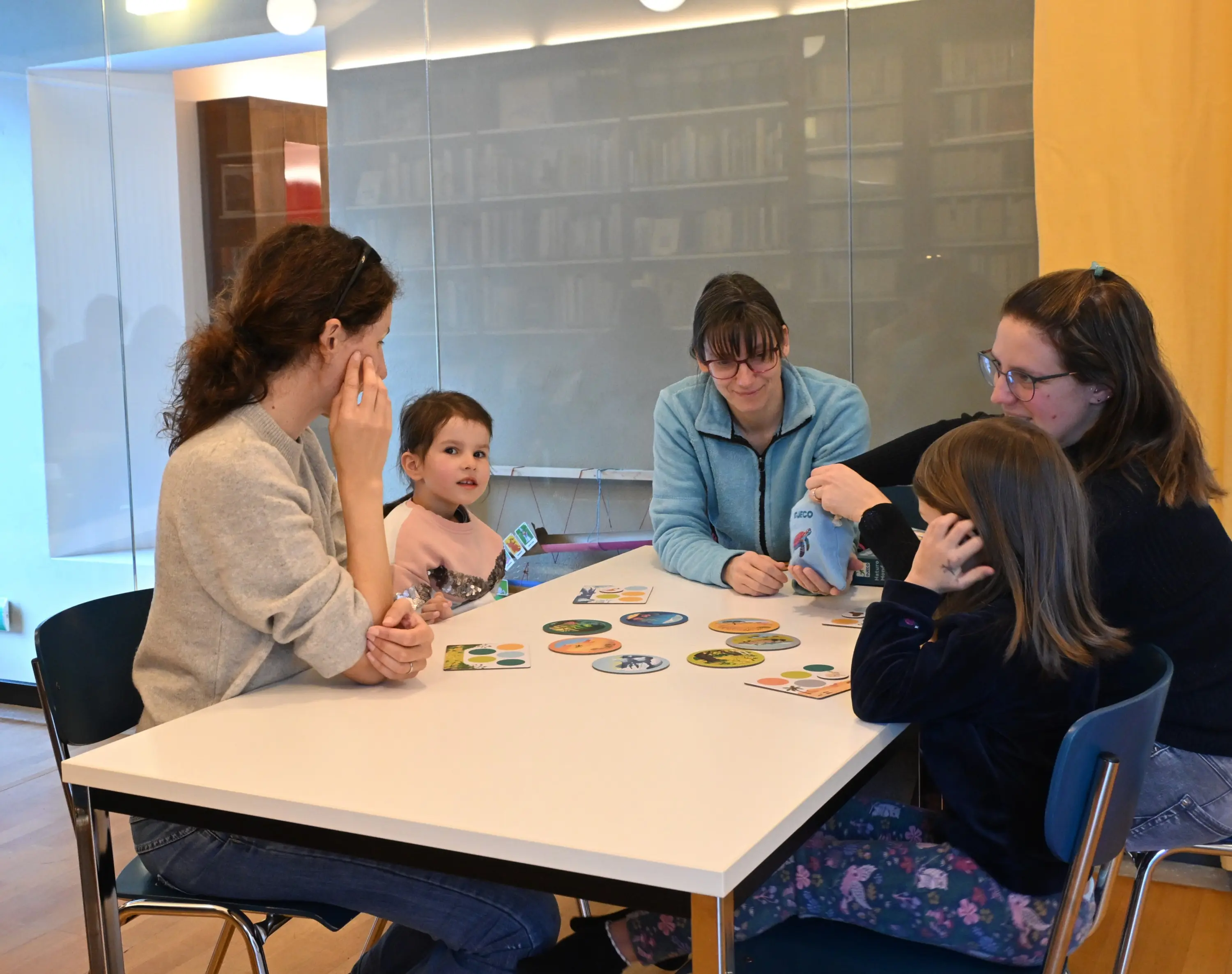 Gruppo di adulti e bambini seduti attorno a un tavolo, impegnati in un gioco di carte. Sul tavolo ci sono vari cerchi colorati con immagini. L'amb