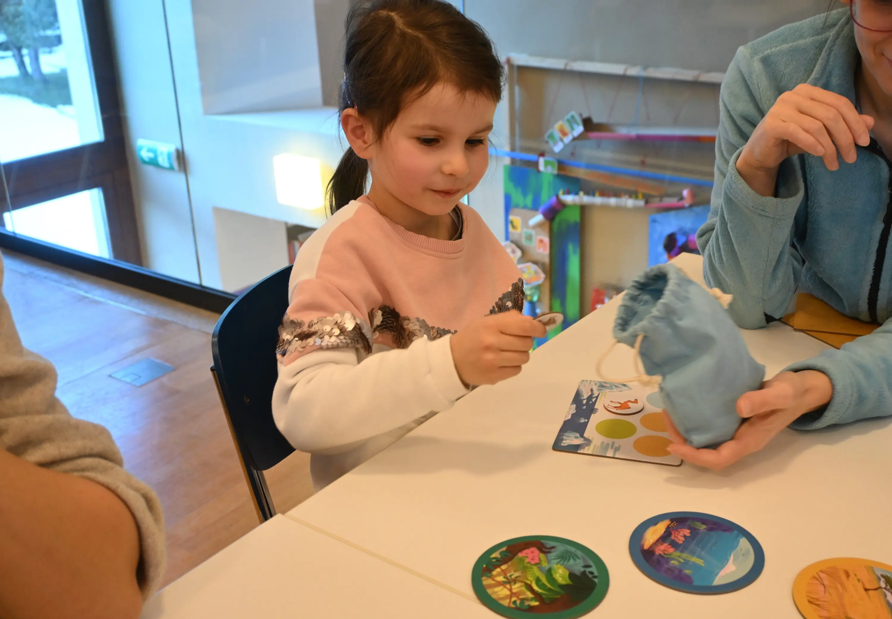 Una bambina sta giocando a un gioco da tavolo, mentre tiene un pezzo di gioco in mano. Un adulto accanto a lei sta mostrando un sacchetto di tessuto. Sul