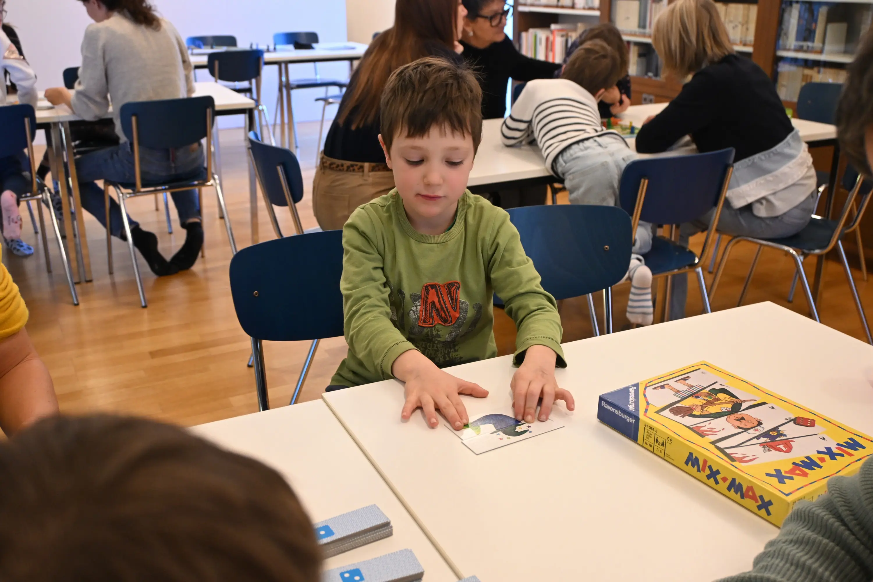 Un bambino con una maglietta verde si concentra mentre tocca un'immagine su un tavolo. Sullo sfondo, altri bambini e adulti sono seduti a tavoli simili,