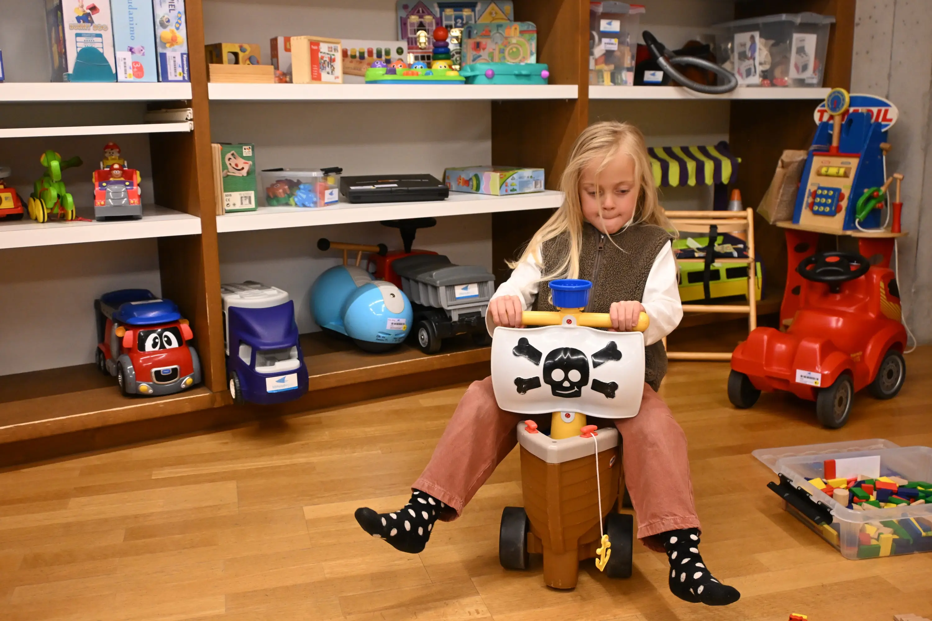 Bambina seduta su un giocattolo a forma di nave pirata, con un teschio disegnato frontalmente, mentre gioca in una stanza piena di giochi e gioc