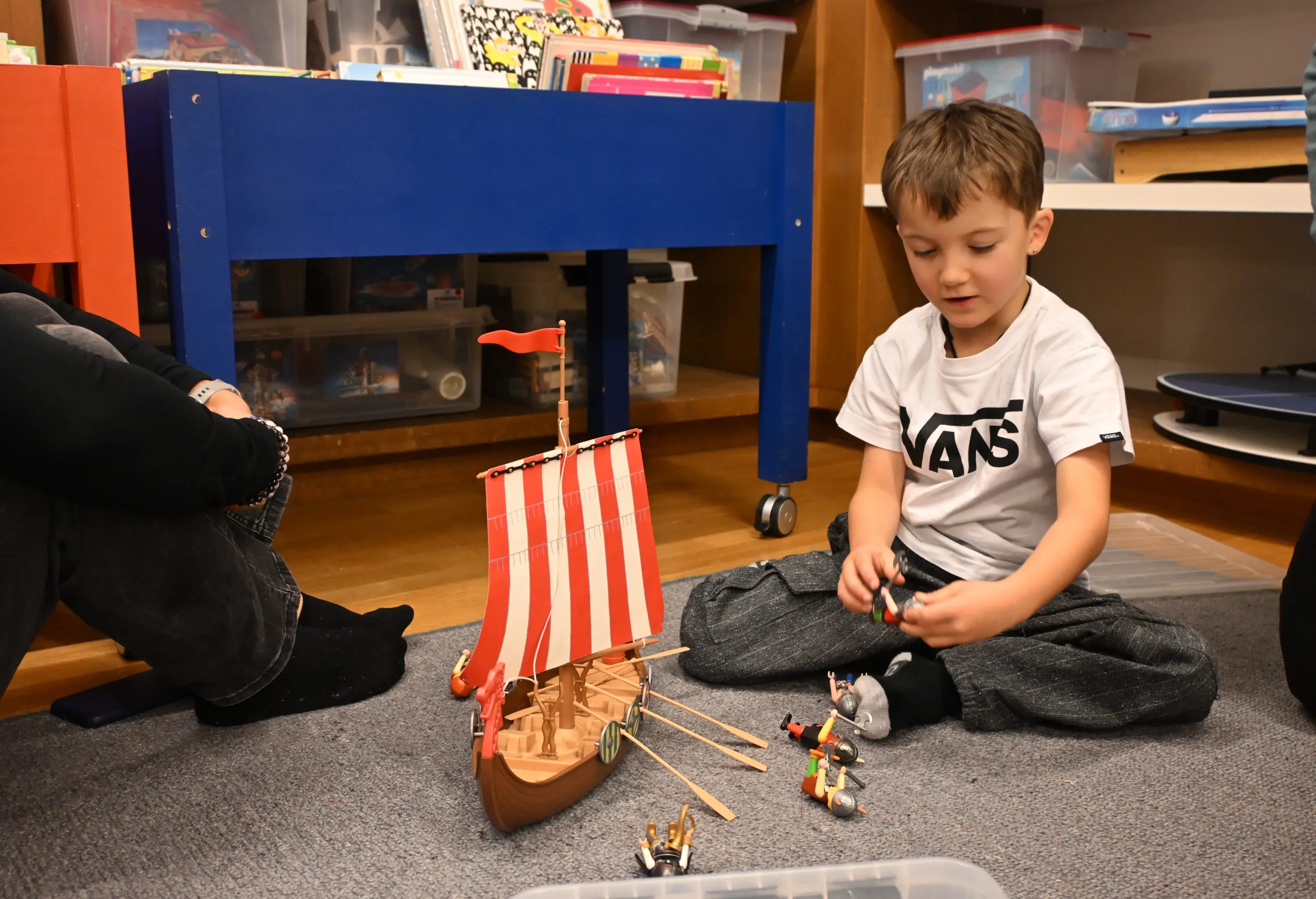 Un bambino gioca a terra con una nave di legno in miniatura, circondato da figure di pirati mentre altri adulti sono seduti vicino a lui.