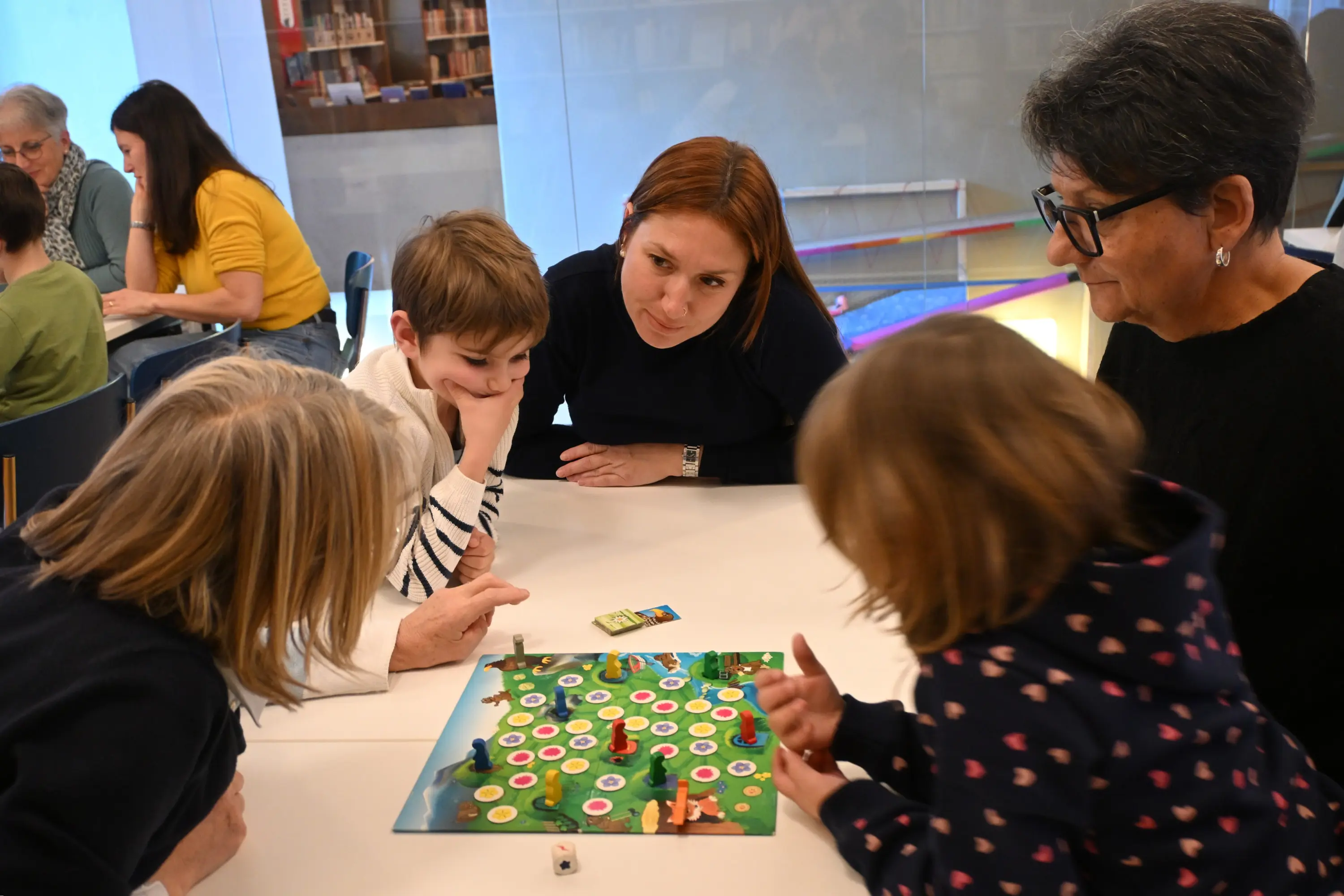 Gruppo di persone, tra cui adulti e bambini, riuniti attorno a un tavolo, intenti a giocare a un gioco da tavolo. Si osservano e discutono