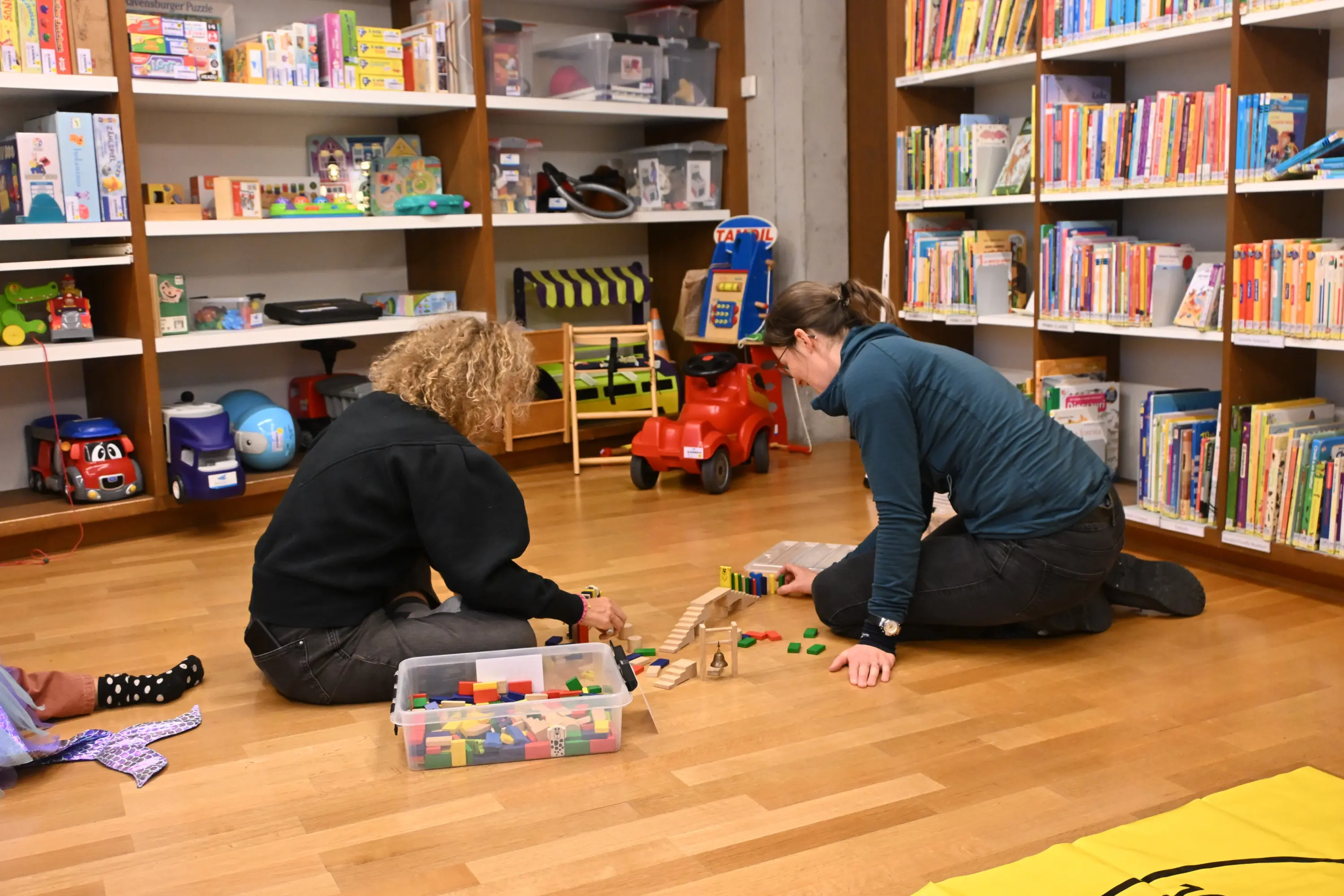 Due persone, sedute sul pavimento di una biblioteca, giocano con dei mattoncini colorati. Intorno a loro, scaffali pieni di libri e giocattoli.