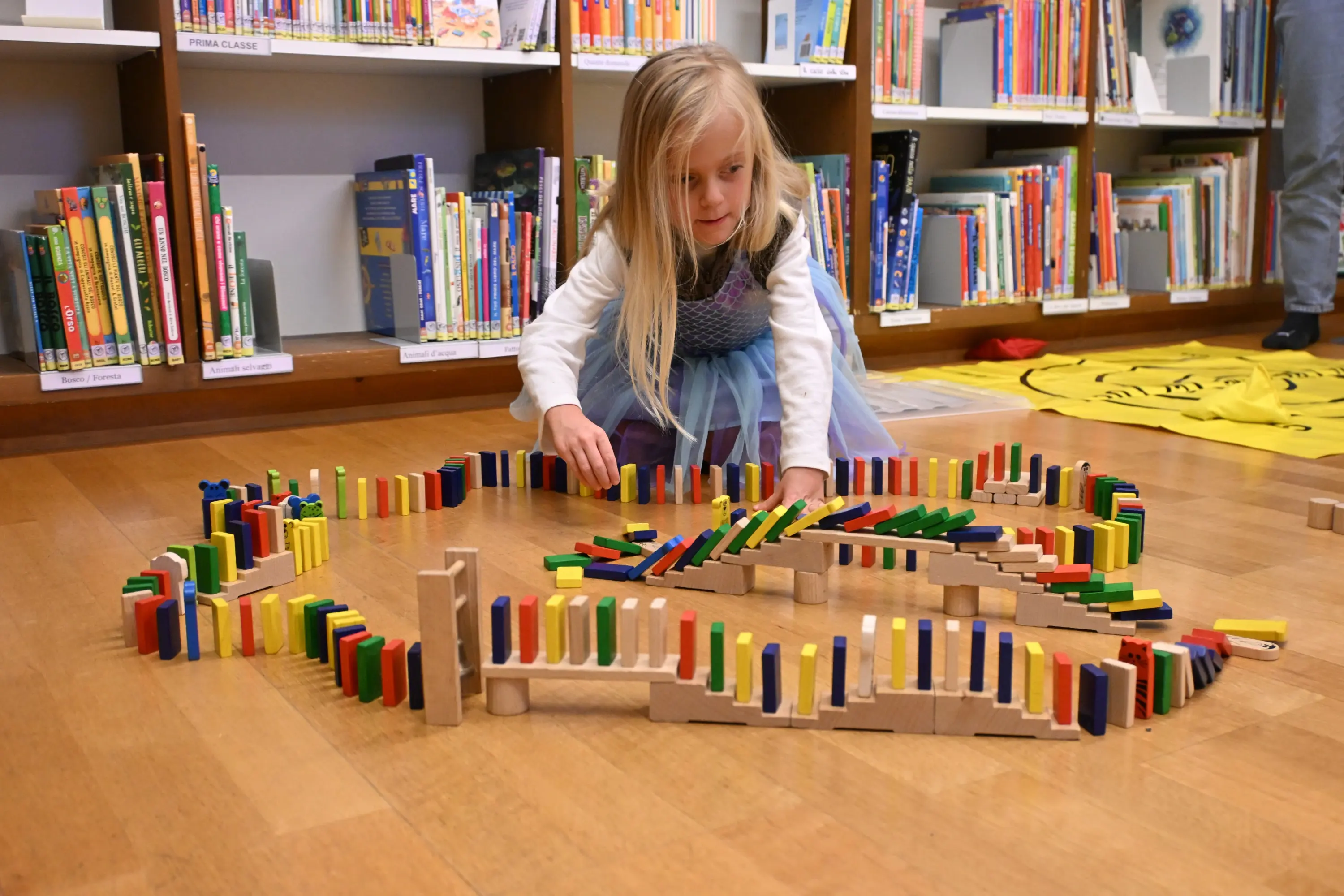 Una bambina sta costruendo una grande struttura di domino colorato in una biblioteca, circondata da scaffali pieni di libri.