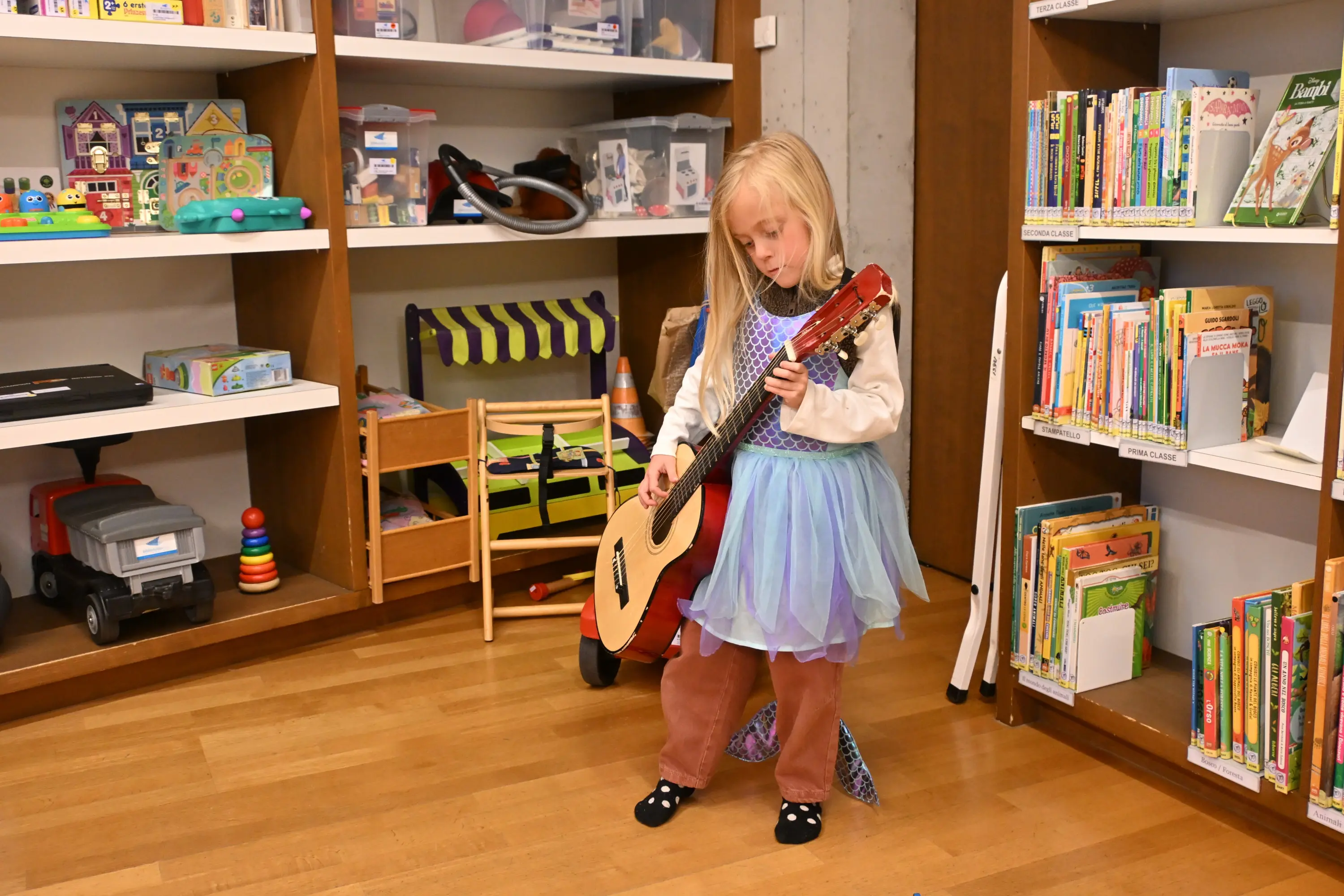 Bambina vestita con un abito colorato che suona una chitarra in una sala giochi, circondata da libri e giocattoli.