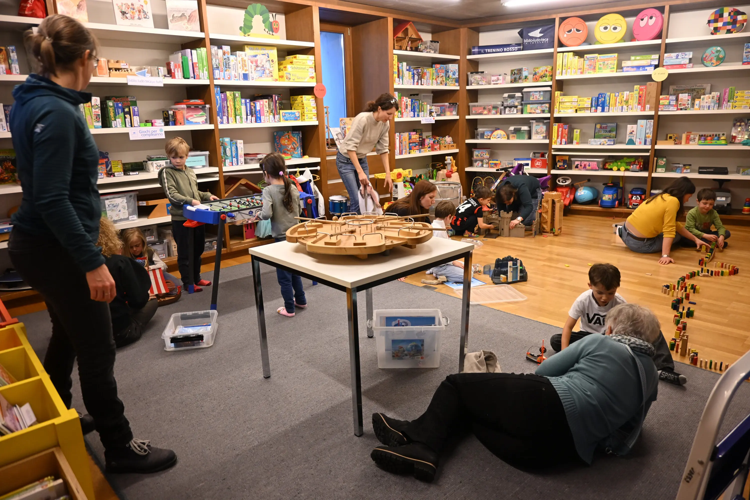Una sala giochi con diverse persone, tra cui bambini che giocano con giochi e costruzioni. Alcuni adulti supervisionano l'attività. Gli scaffali sono pieni di libri e giochi colorati