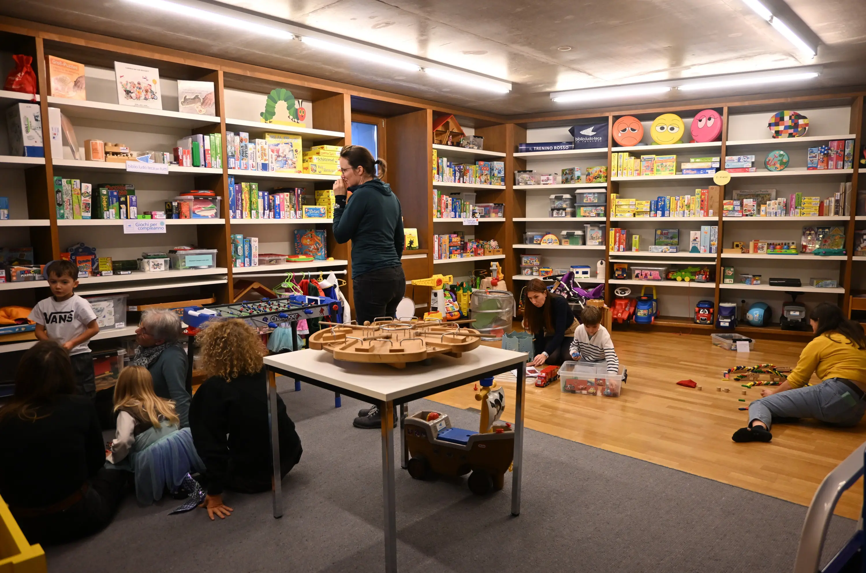 Una stanza di gioco colorata con bambini che giocano a diverse attività. Una donna si occupa di un gruppo di bambini mentre altri giocano a terra. Gli scaffali sono pieni di giochi e