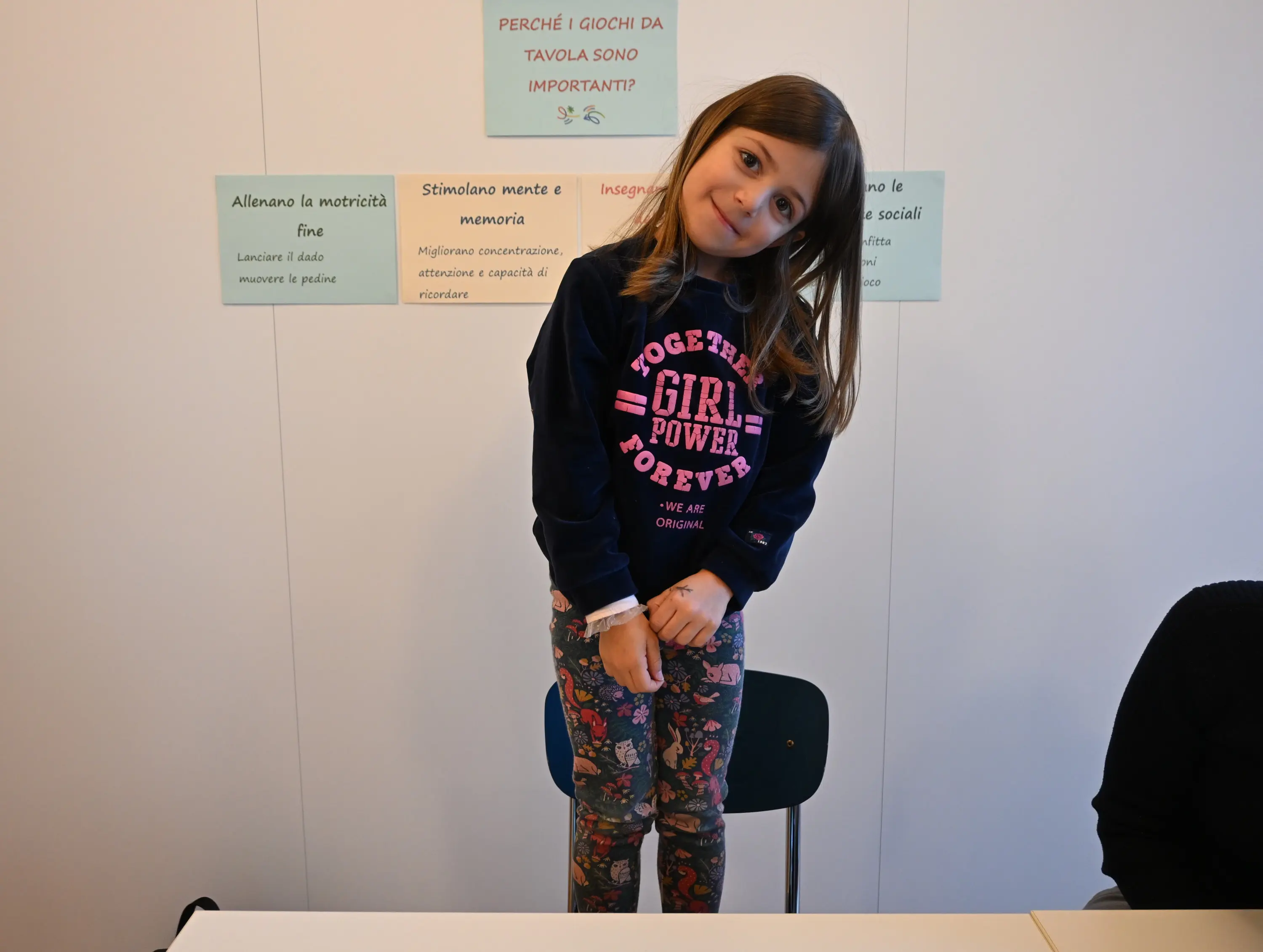 Bambina con maglione blu scuro e pantaloni con stampa colorata, in piedi accanto a un tavolo. Sorridente, inclina leggermente la testa con le mani