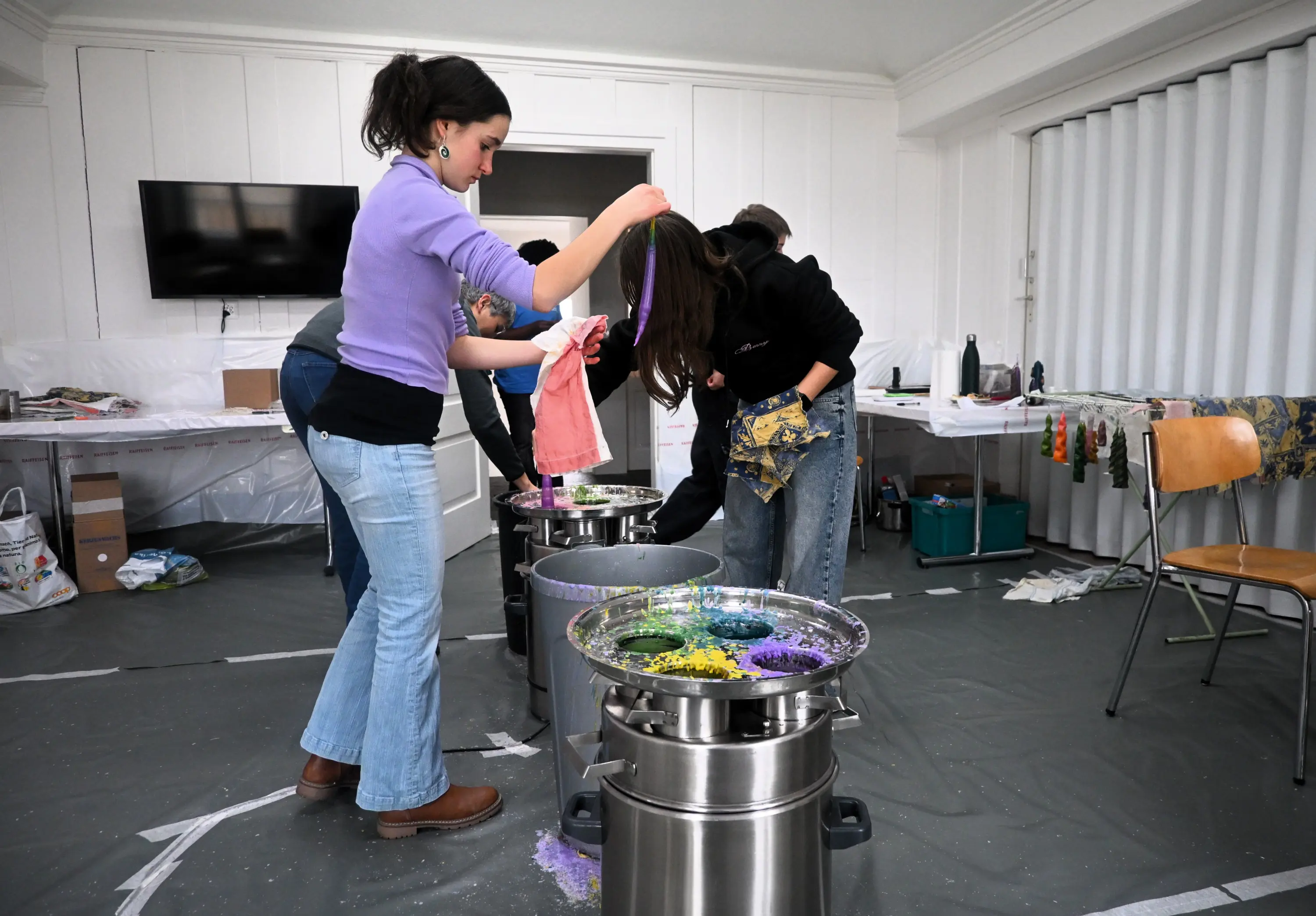 Gruppo di persone che lavorano insieme in una stanza, mentre applicano colori su stoffe utilizzando contenitori metallici. Alcuni partecipanti indossano guanti e Manichini color