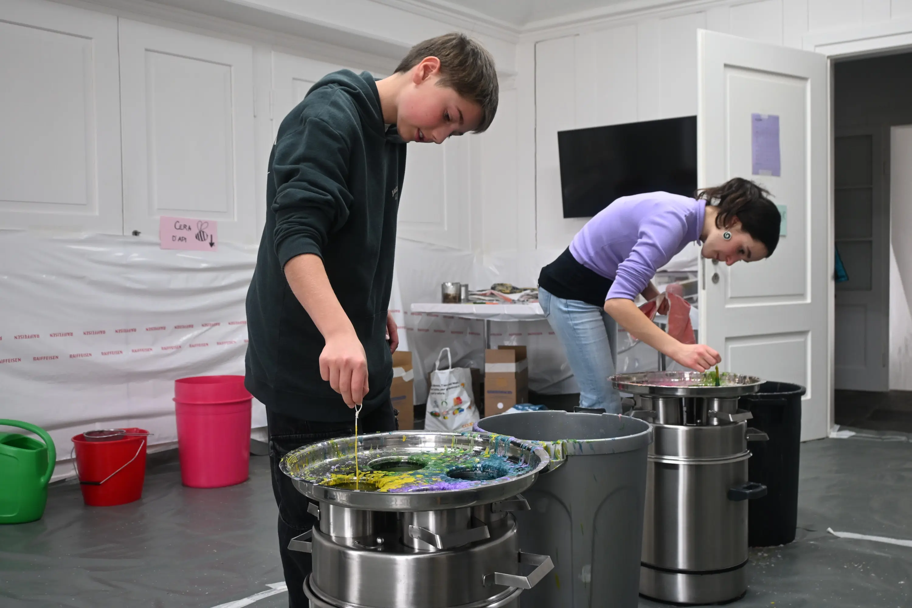 Un ragazzo mescola colori vivaci in un contenitore, mentre una giovane donna lavora su un altro progetto alle sue spalle. Sono in una stanza ben illuminata, circondati da