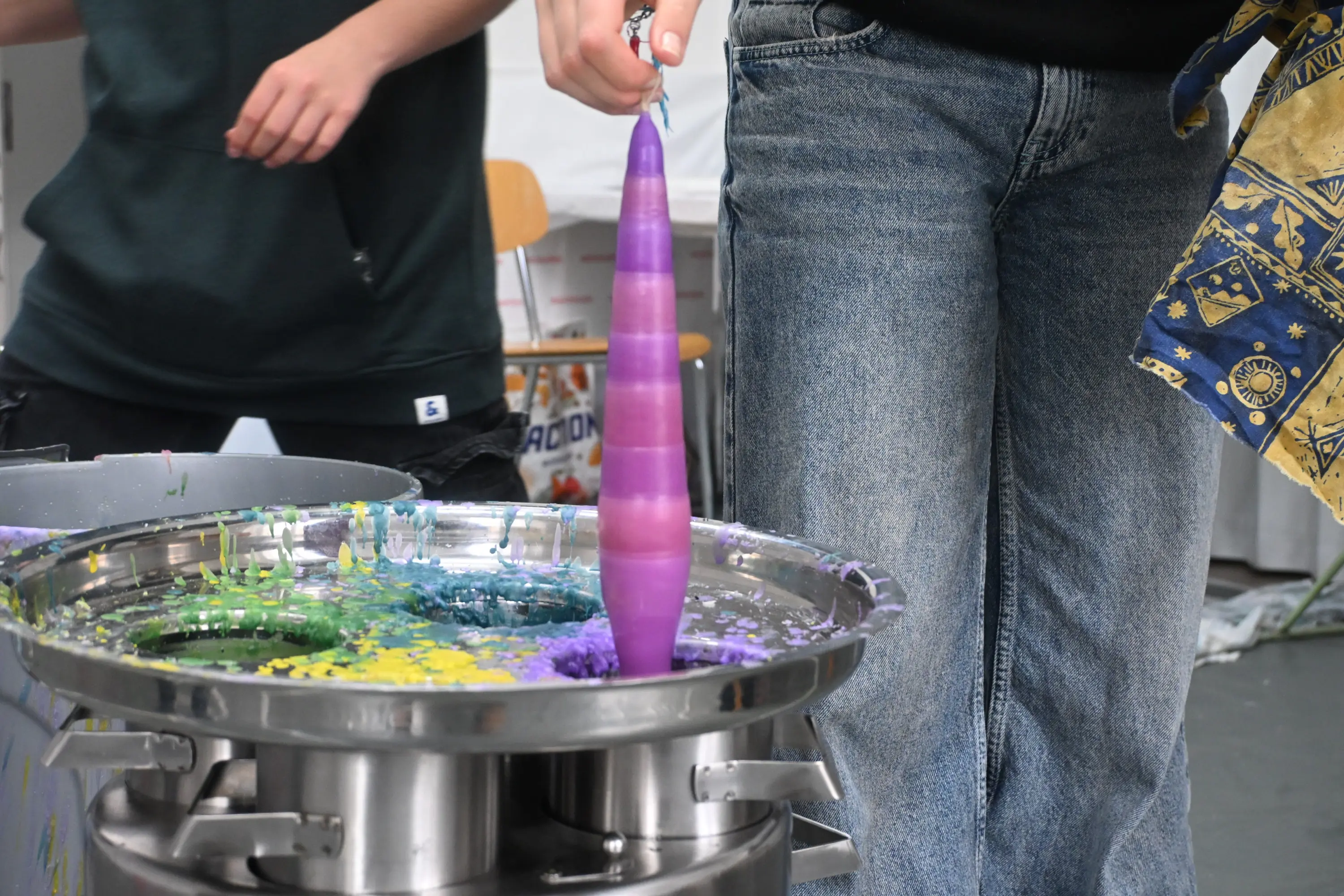 Un bastone colorato viene immerso in un contenitore di vernice, creando effetti artistici. Sono visibili due persone, una delle quali indossa un pantalone di jeans.