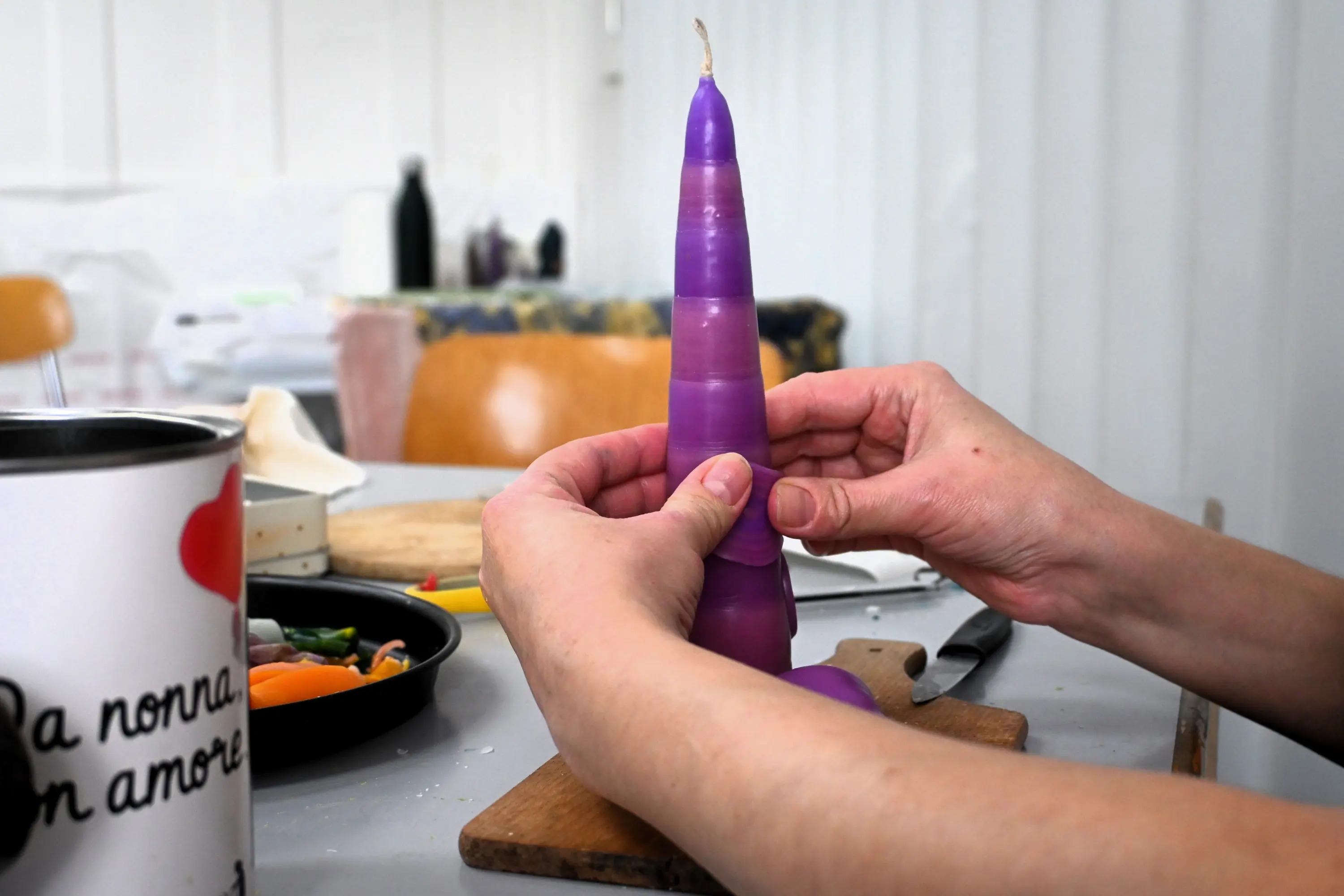 Due mani modellano una candela viola a forma conica su un piano di lavoro. Sullo sfondo si intravedono utensili da cucina e una sedia.