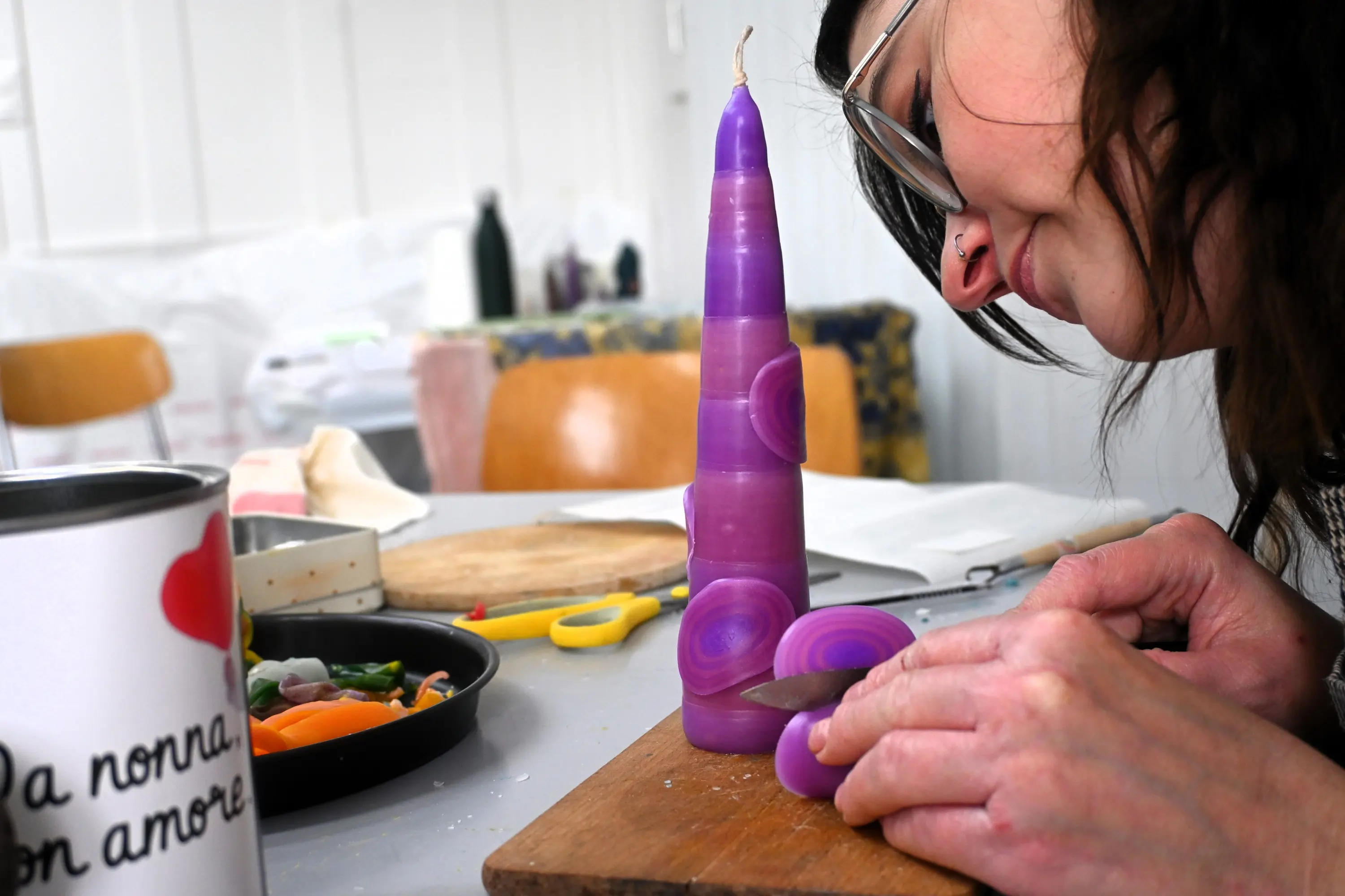 Una donna sta intagliando una candela di cera viola su un tagliere di legno, con un coltello. Sul tavolo ci sono anche verdure e altri materiali da lavoro