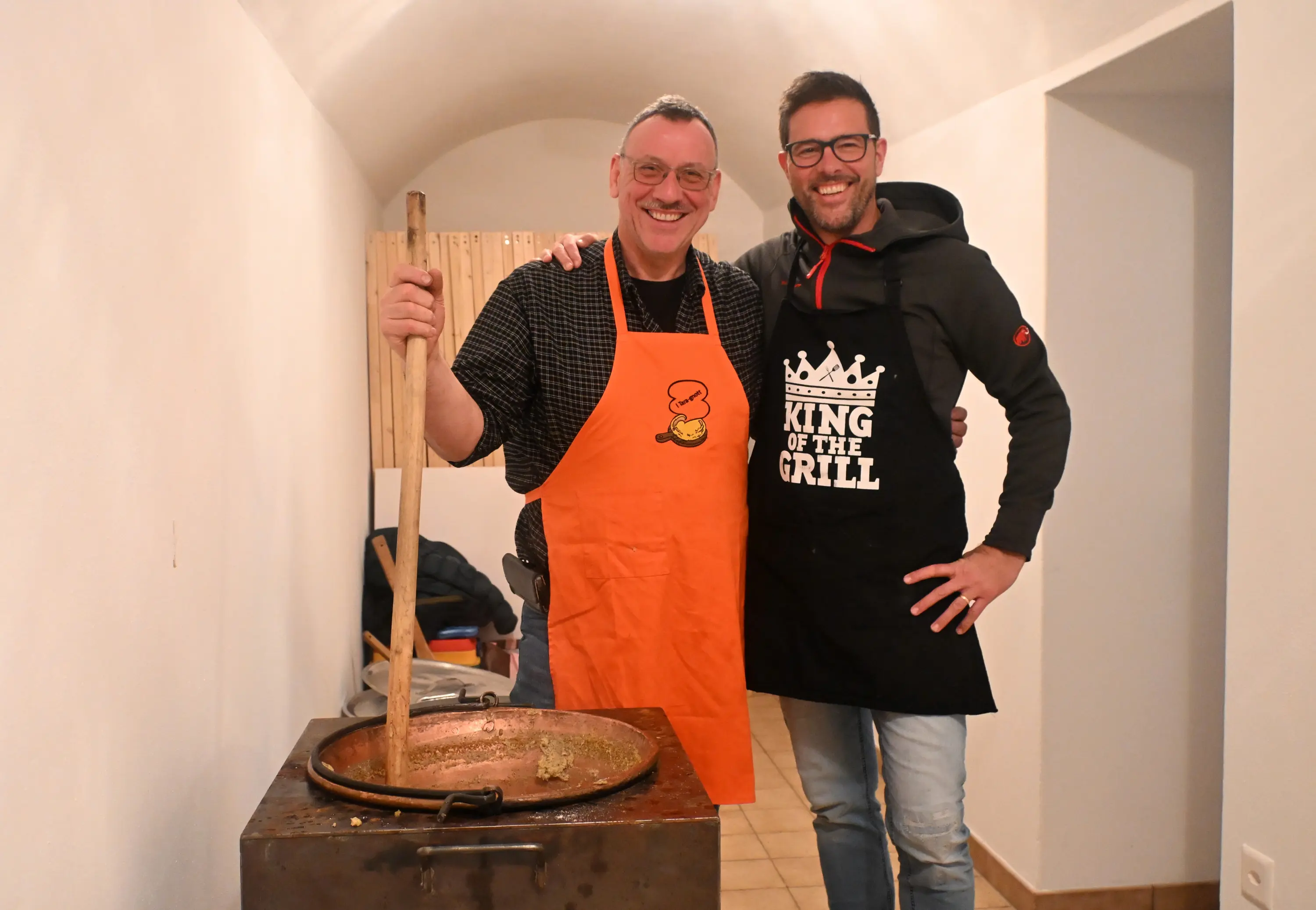 Due uomini sorridenti in cucina, uno indossa un grembiule arancione e l'altro un grembiule nero con la scritta "KING OF THE GRILL". Stanno entr