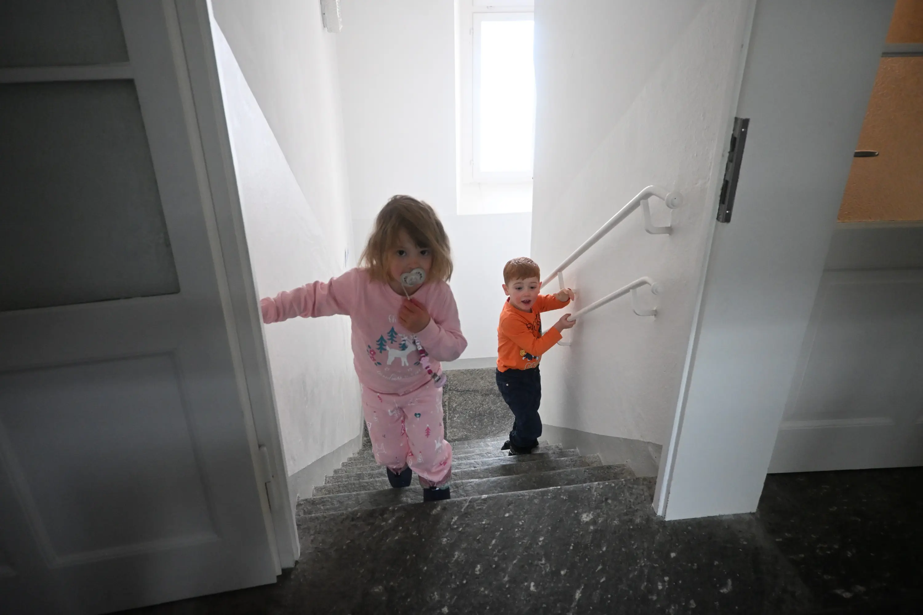 Due bambini stanno salendo una scala. La ragazza, con un ciuccio in bocca, indossa un pigiama rosa con disegni di alberi. Il ragazzo,