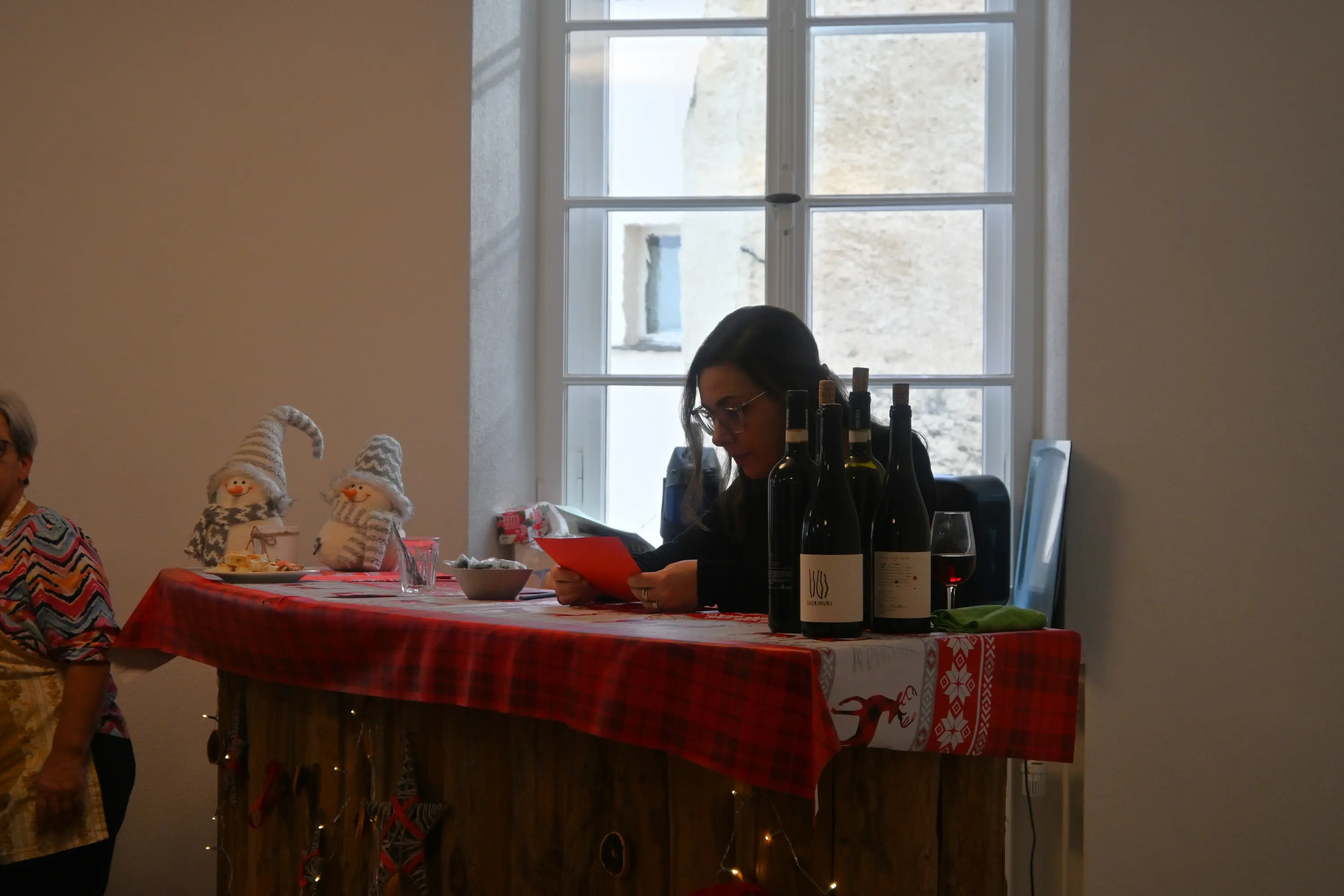 Una donna seduta al bancone con una tavola decorata per Natale, mentre legge un documento. Sul bancone ci sono bottiglie di vino e decorazioni natalizie. Sfondo luminoso