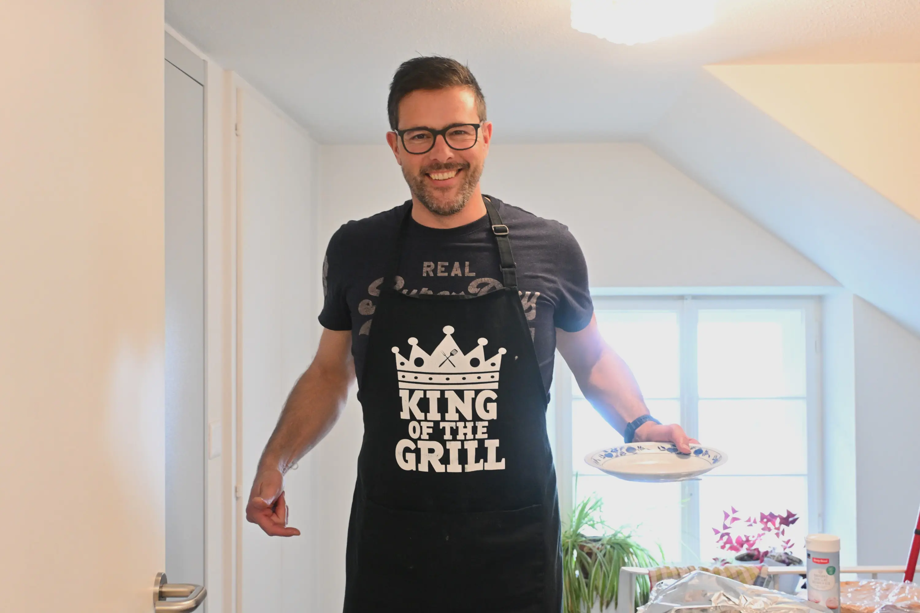 Un uomo sorridente indossa un grembiule nero con la scritta "King of the Grill" mentre tiene un piatto in mano. È in una cucina con una finestra che illumina