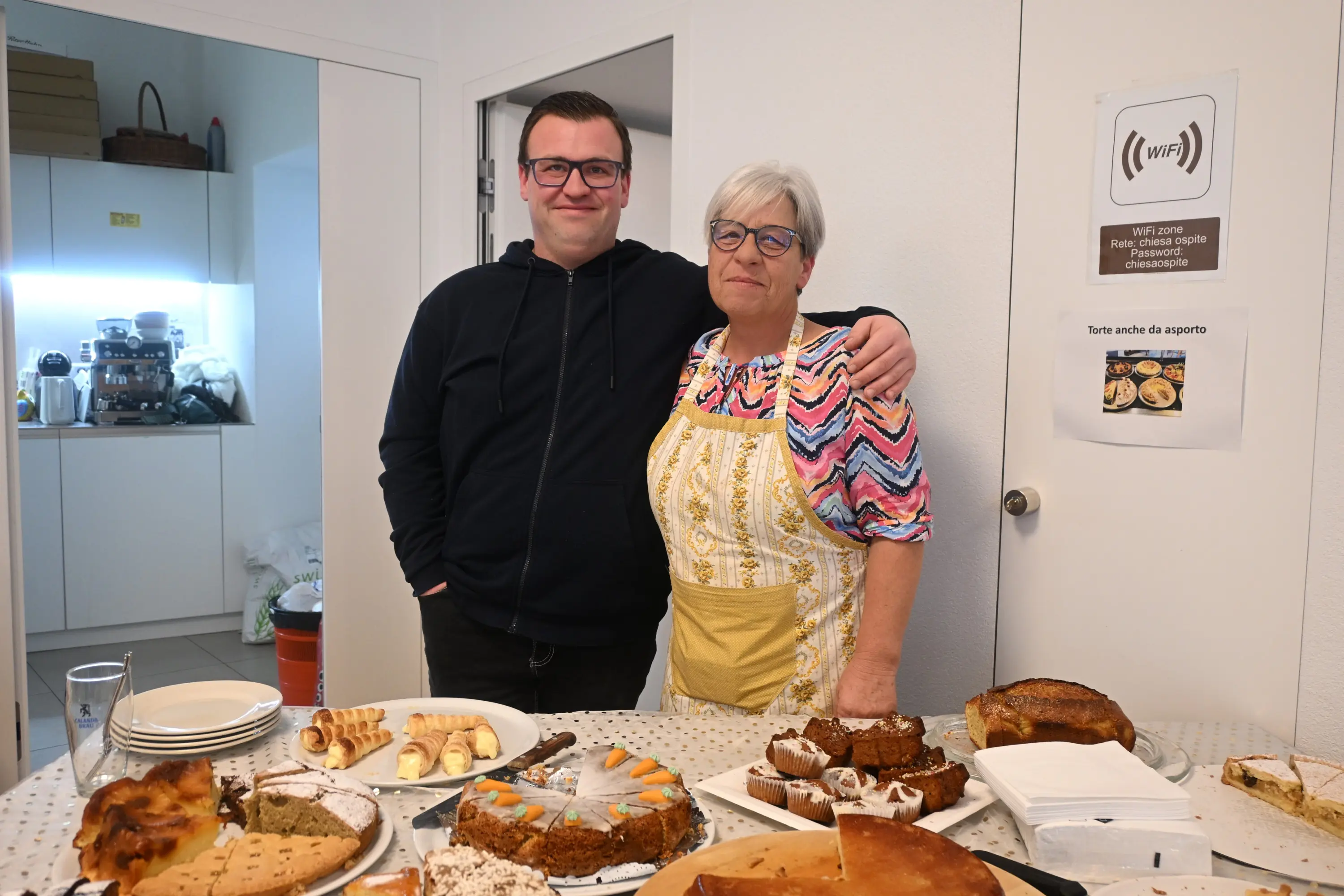 Due persone sorridenti, un uomo giovane e una donna di mezza età, posano insieme davanti a un tavolo imbandito con vari dolci e stuzzichini. La donna