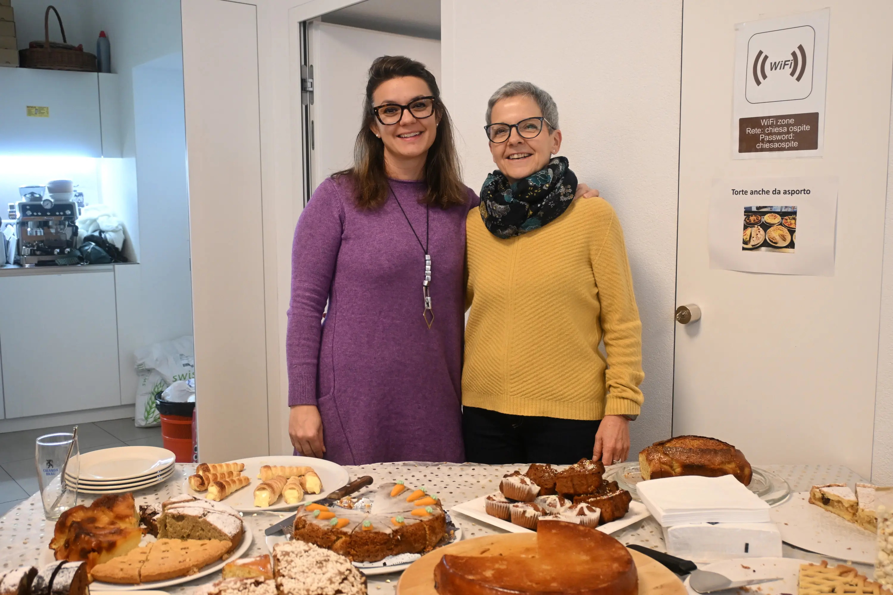 Due donne sorridenti posano insieme davanti a un tavolo imbandito con diversi dolci e piatti. Una porta bianca si vede sullo sfondo.