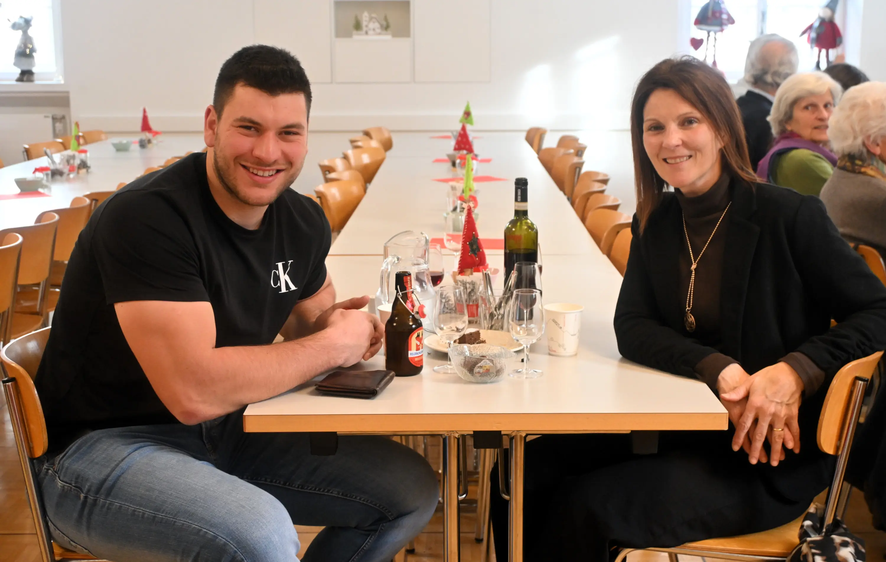 Due persone sorridenti sedute a un tavolo imbandito in una sala decorata per le festività. L'uomo indossa una maglietta nera e ha un bicchiere