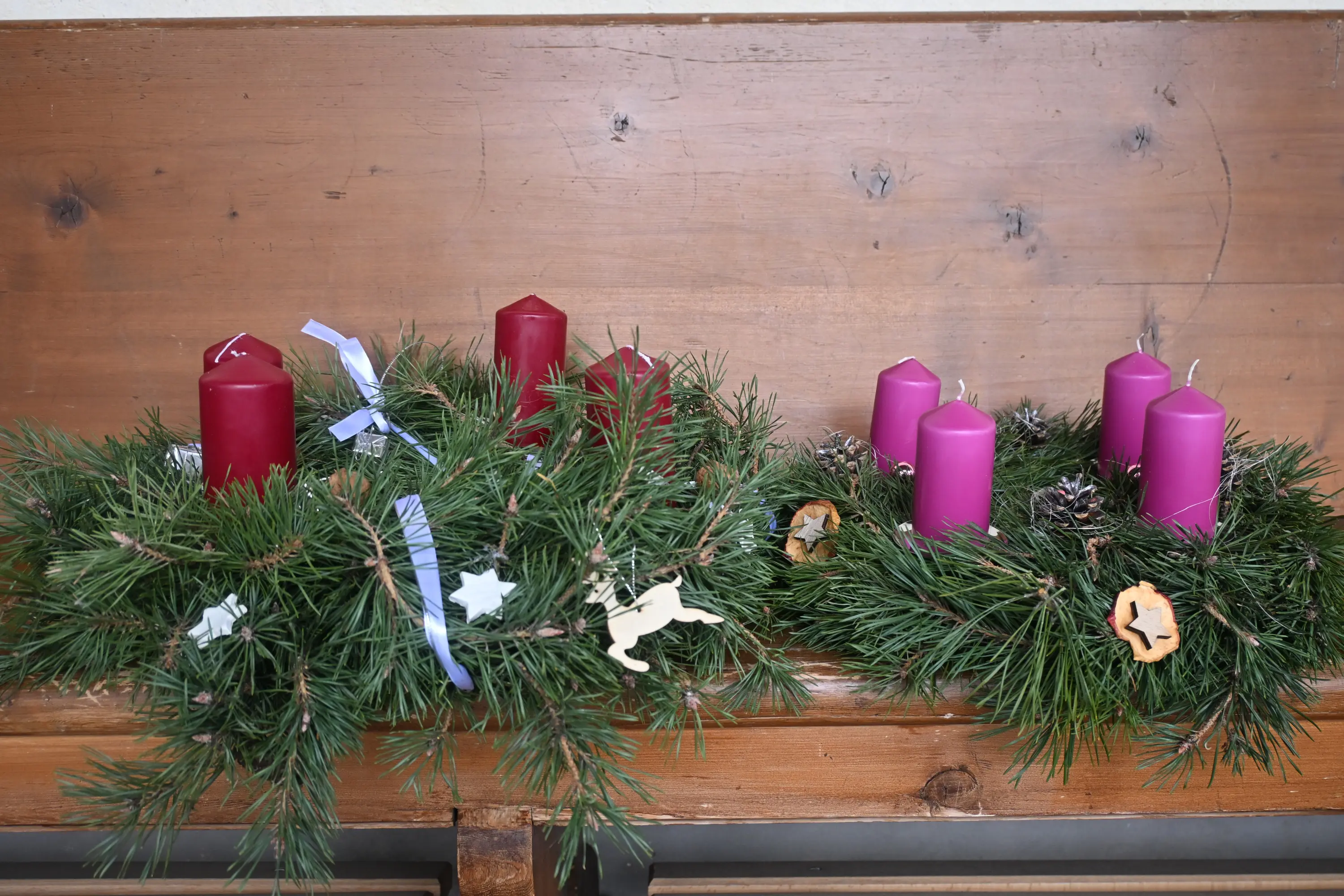 Corone di avvento con candele colorate in tonalità rosse e viola, decorate con nastri e ornamenti, posizionate su un tavolo di legno.