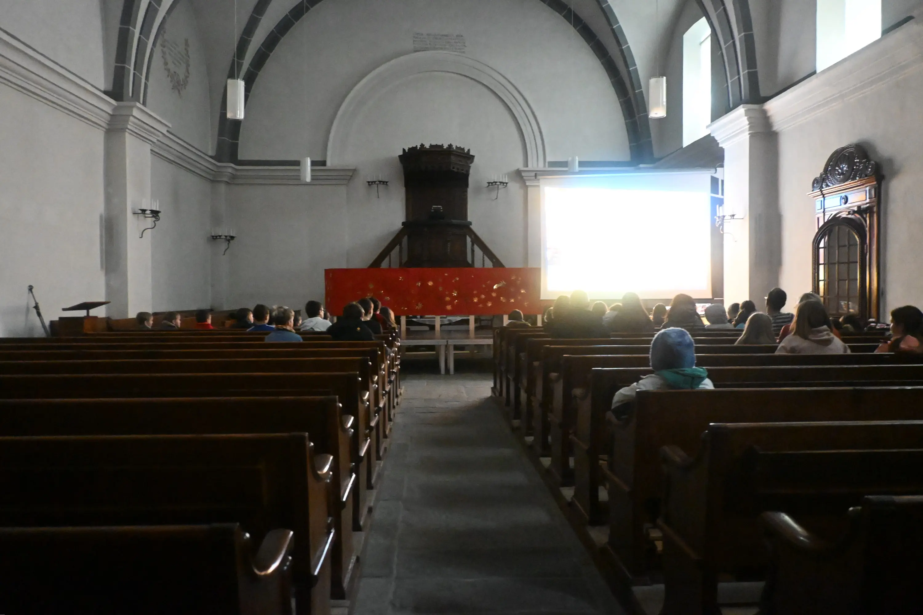 Interno di una sala per conferenze con panche di legno. Persone sedute di fronte a uno schermo proiettore, illuminato da una luce tenue. Sullo sfondo