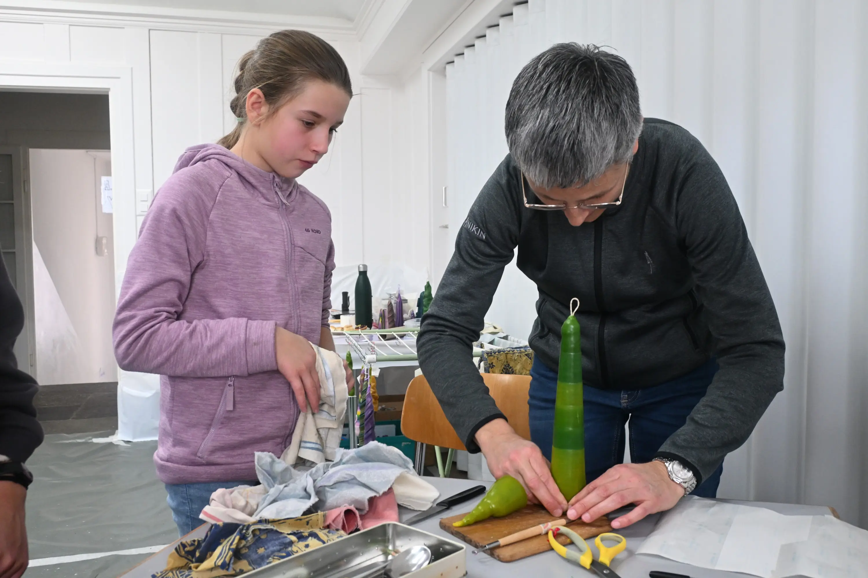 Una donna adulta sta realizzando una candela verde con una ragazza giovane che osserva attentamente. Entrambi sono all'interno di una stanza ben illuminata, circondati da materiali