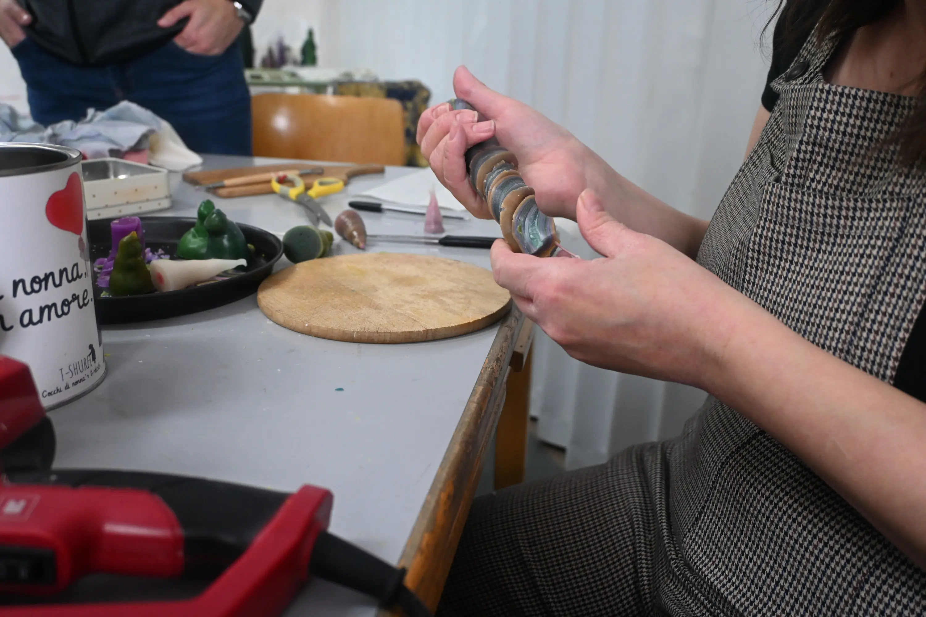 Una persona tiene in mano una serie di pietre colorate su un tavolo da lavoro. Sul tavolo ci sono strumenti come forbici e una base di legno. Un'altra persona è par