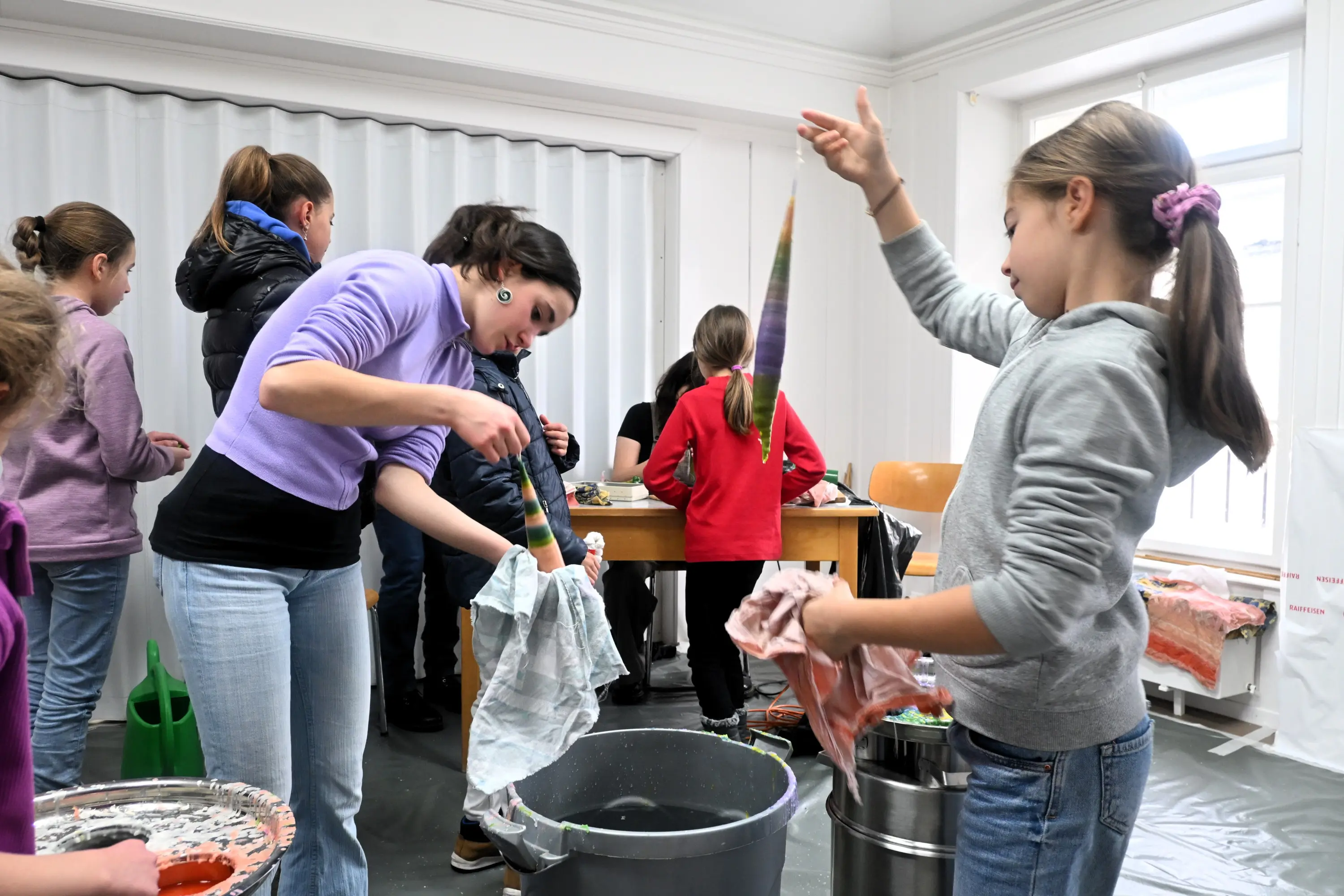 Un gruppo di bambini e adulti è impegnato in un'attività di tintura, con una donna che immerge un tessuto in un contenitore e una ragazza che tiene un altro