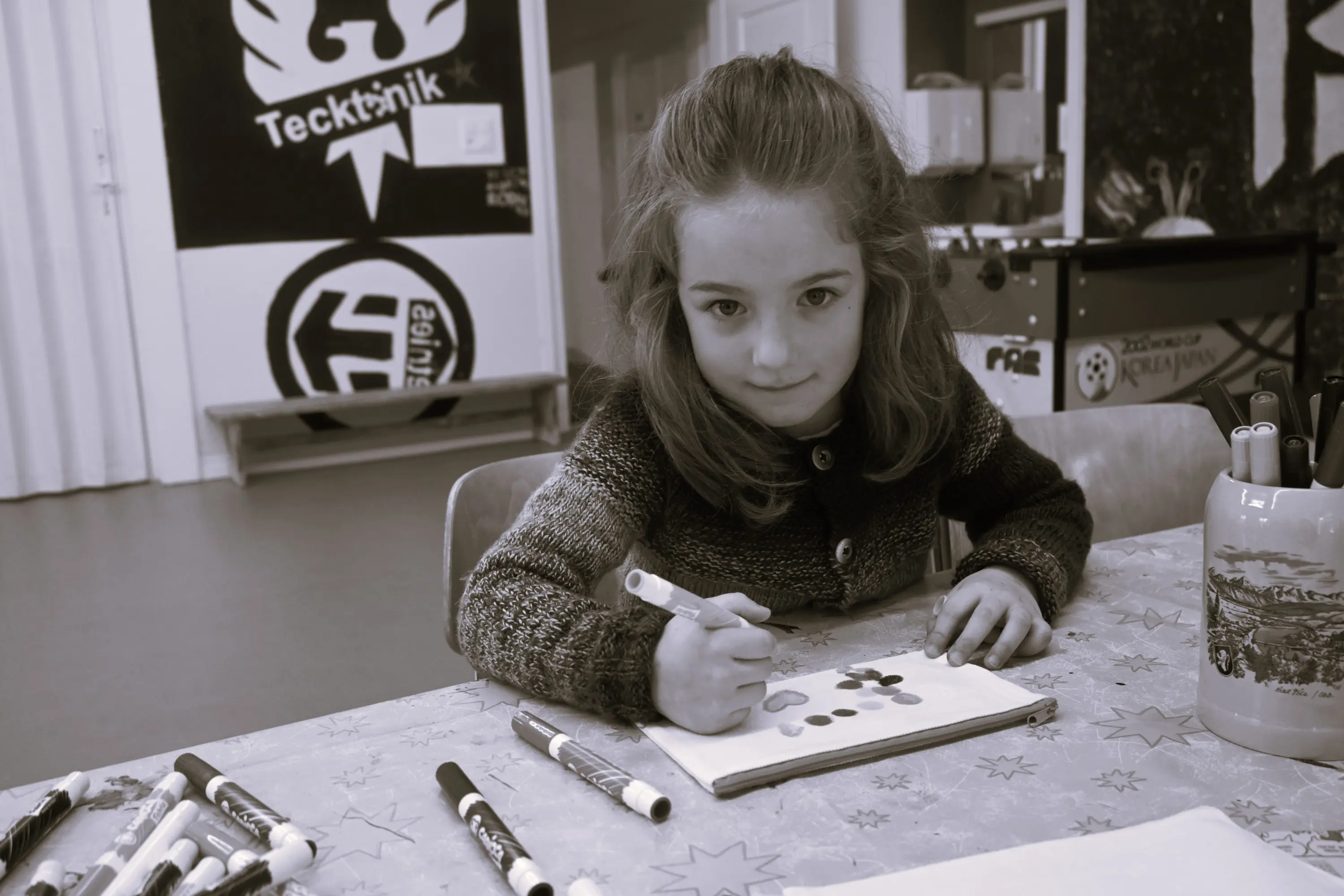 Una bambina disegna su un foglio con un pennarello, seduta a un tavolo. Sullo sfondo ci sono colori e attrezzature artistiche. La scena è in