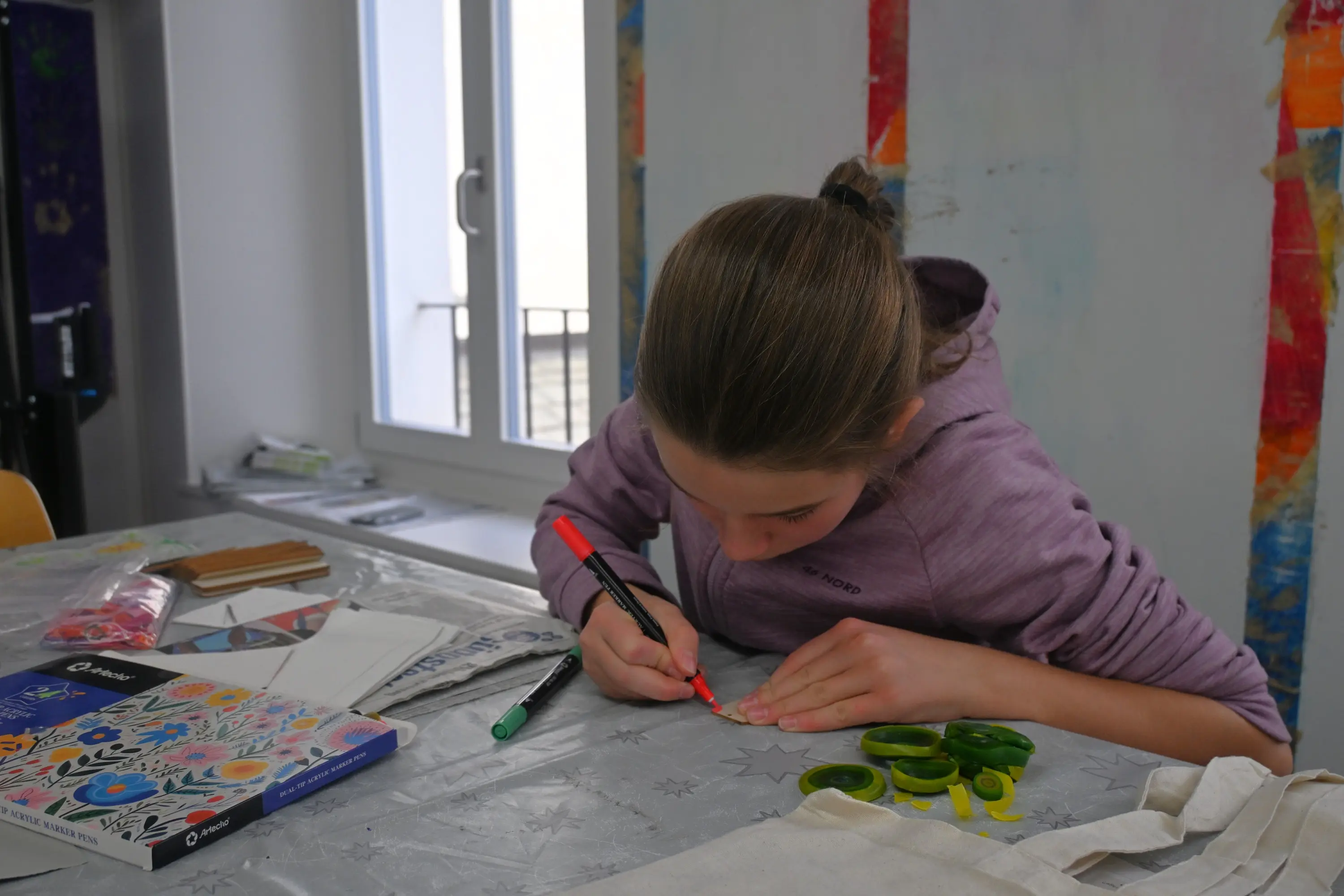 Una ragazza sta colorando con un pennarello su un tavolo di lavoro. Ha dei materiali artistici sparsi intorno e mostra concentrazione. La luce entra da una finestra vic